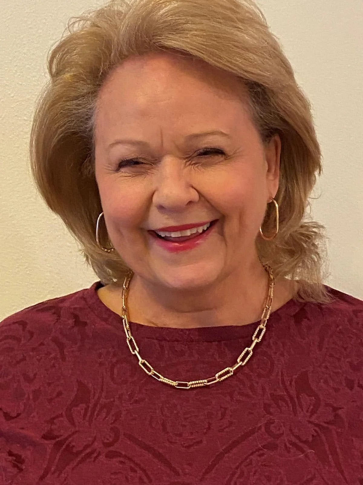 A smiling older woman with blonde hair, wearing gold hoop earrings, a gold chain necklace, and a red textured top, standing against a plain background.