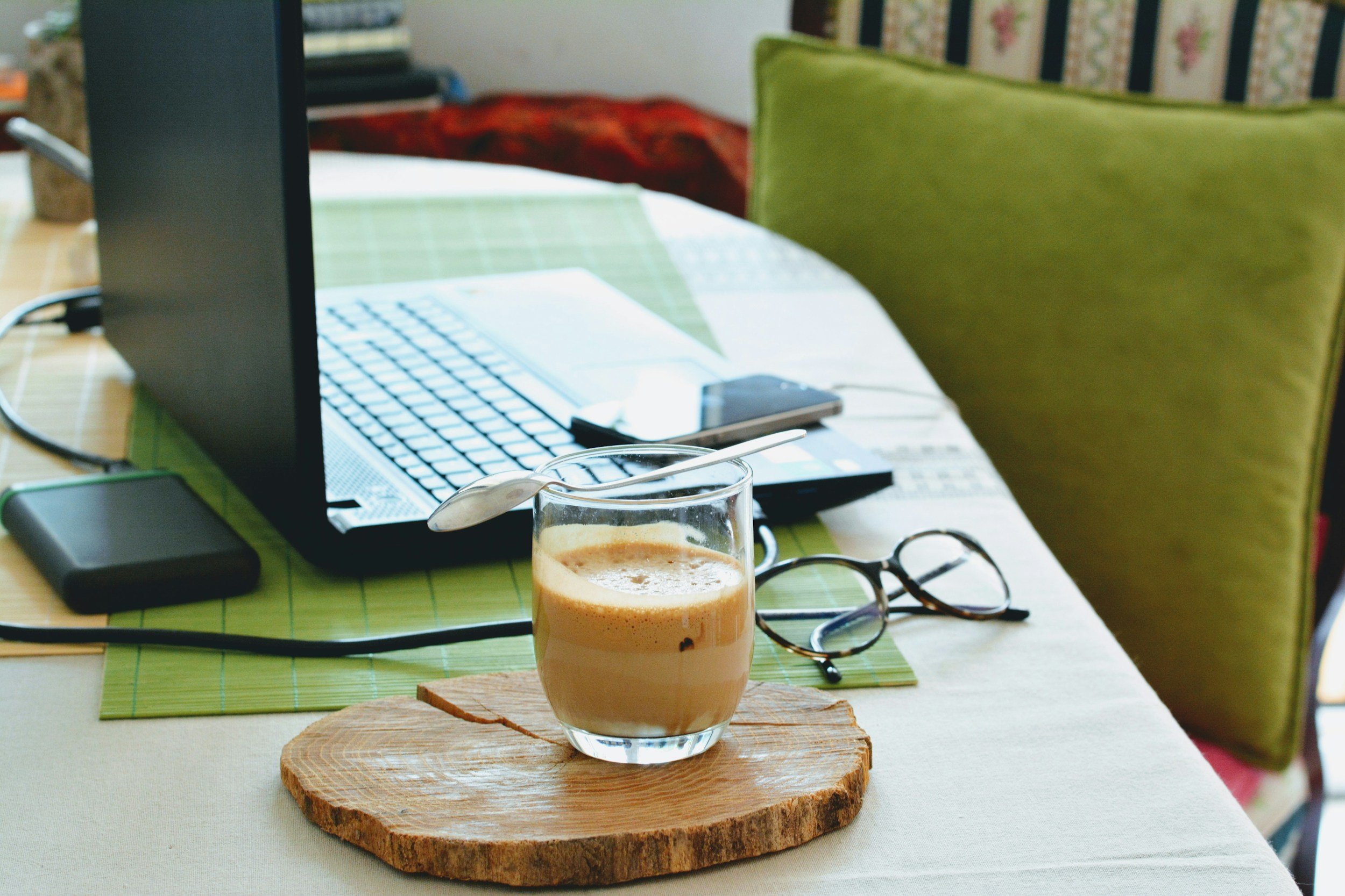 A dining table with a laptop, glasses, a smartphone, and a glass of coffee on a wooden coaster.