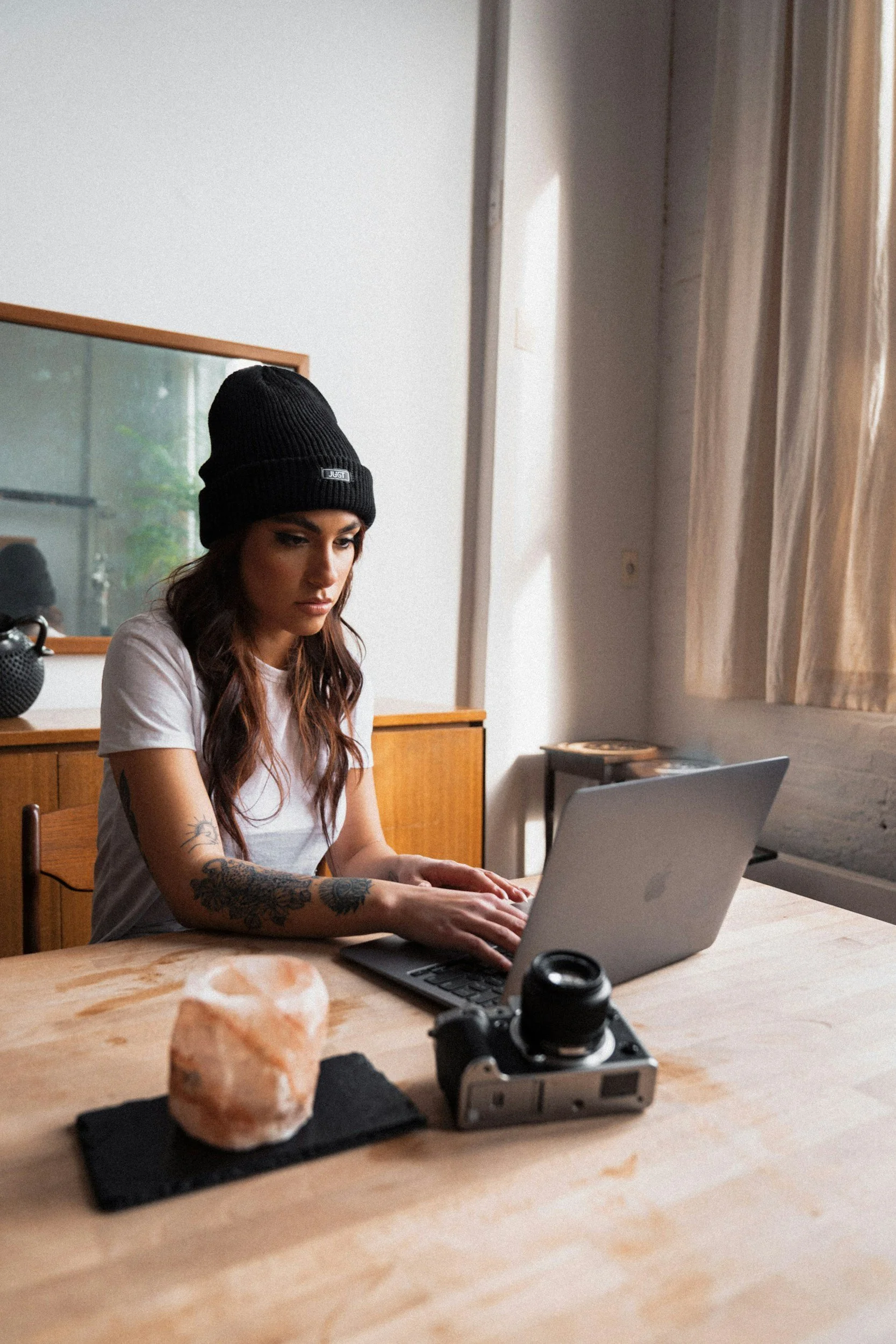 A young woman with tattoos on her arms sitting at a wooden table, wearing a black knit beanie and a white t-shirt, working on a silver laptop. There is a camera and salt lamp on the table, and a mirror, window, and curtain in the background.