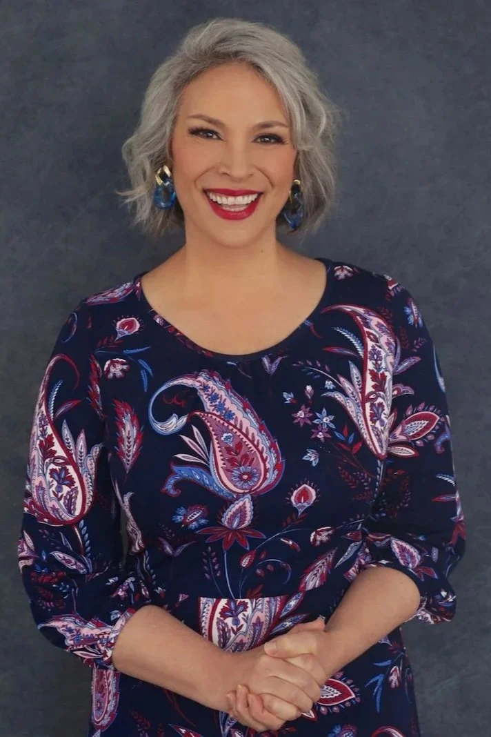 A woman with shoulder-length gray hair, wearing makeup and red lipstick, smiling, dressed in a dark blue dress with a colorful paisley pattern, standing in front of a dark gray background.