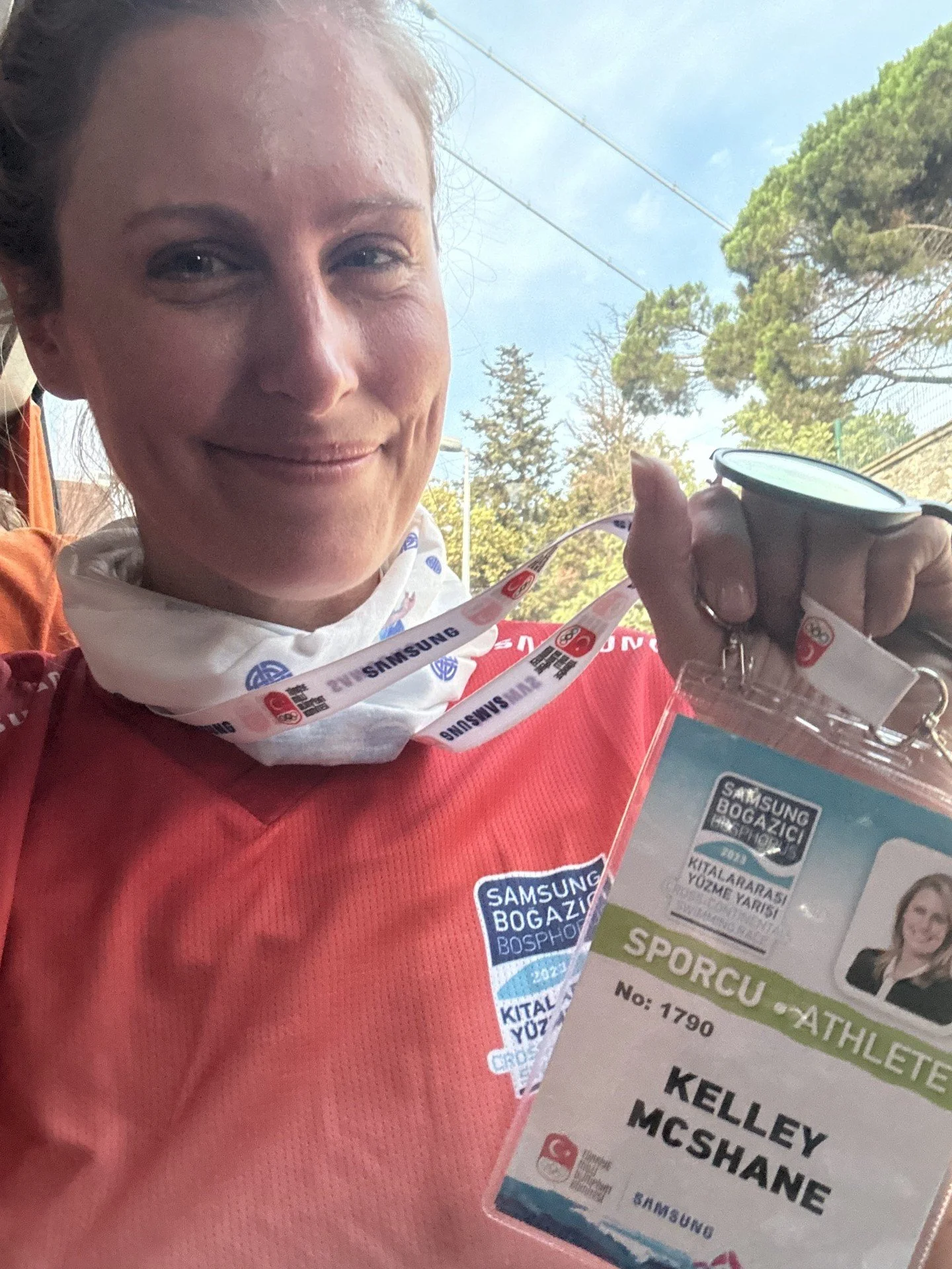 A woman smiling while holding a medal and wearing an orange sports shirt with a badge that reads 'Samsung BOGAZICI BOSPHORUS 2022'. She has a white neck gaiter with logos, and appears to be at an outdoor event with trees and sky in the background.