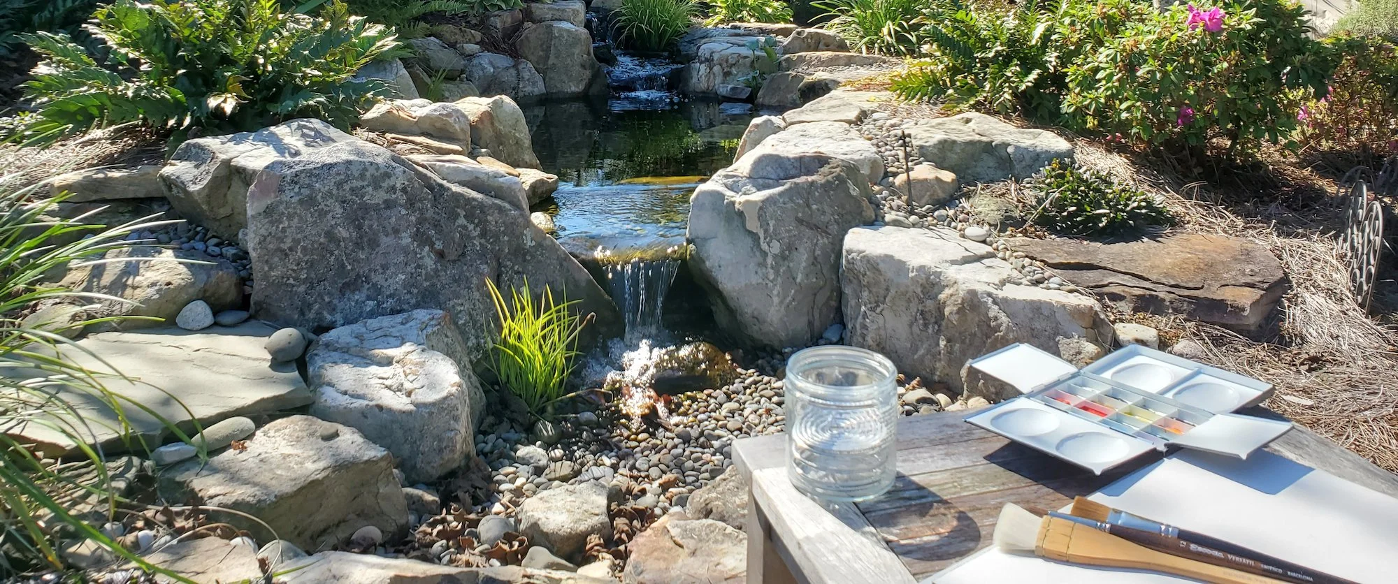 Outdoor watercolor painting setup by a garden stream with rocks, plants, flowing water, a jar of rinse water, open paint palette, brushes, and sketch paper, illustrating Beverly G. McCarter's return to art and exploration of sense of place.
