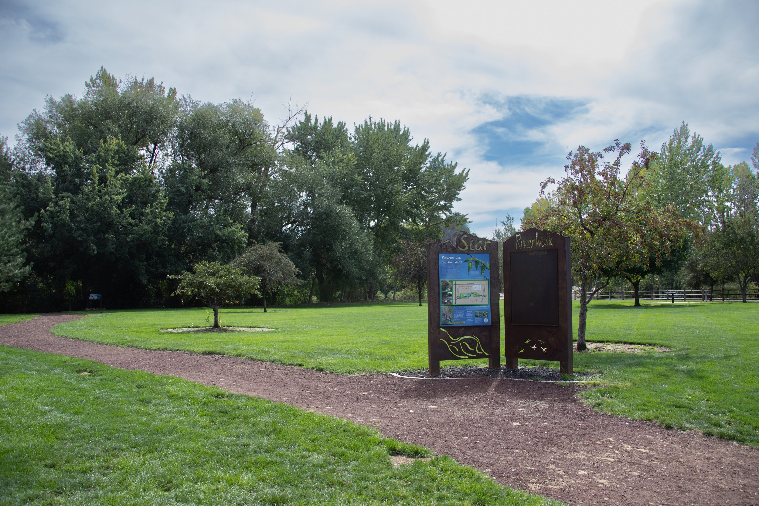Signage in a park with green grass, trees, and a gravel walking path, under a partly cloudy sky.