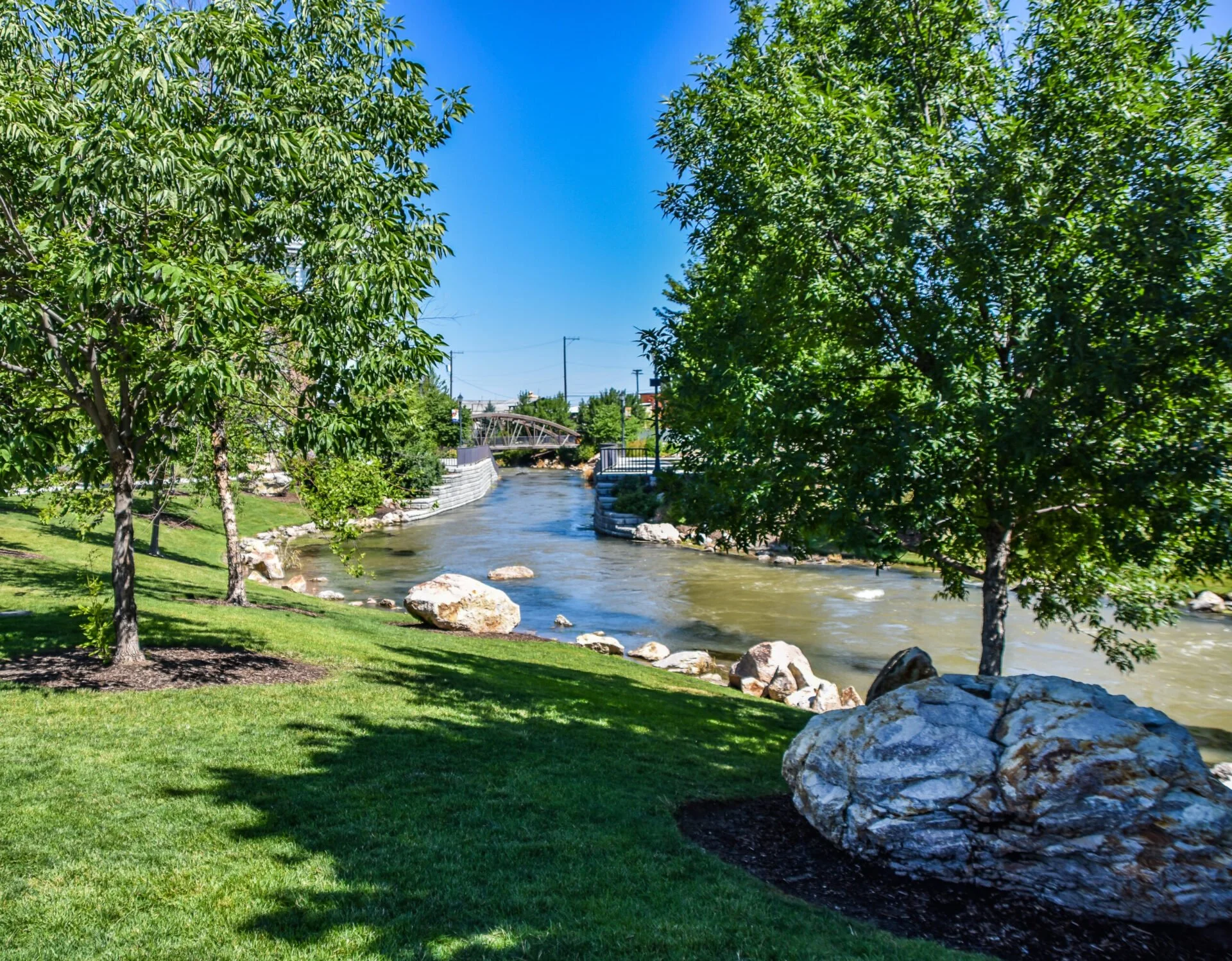 A scenic park with green grass, trees, and a flowing river under a clear blue sky.