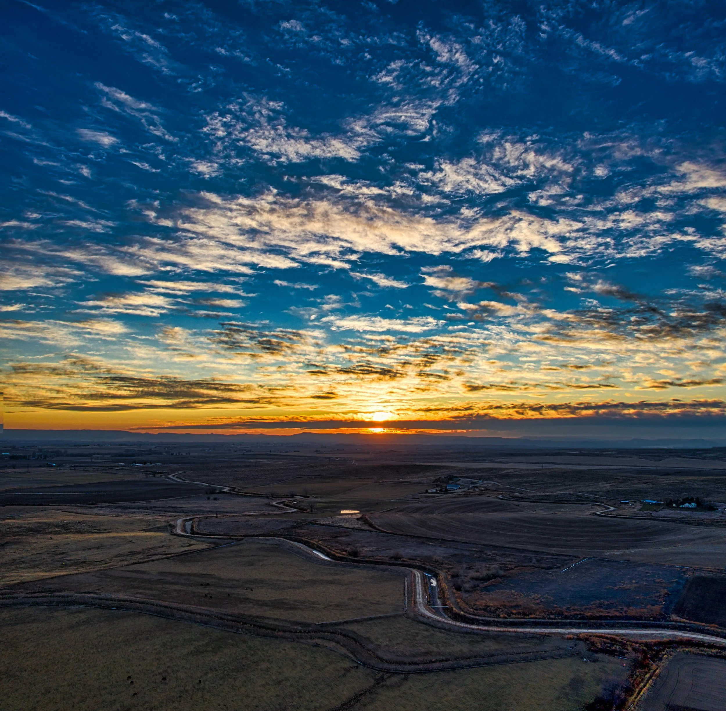 Sunset over rural landscape with winding roads and scattered buildings, partly cloudy sky with rays of sunlight