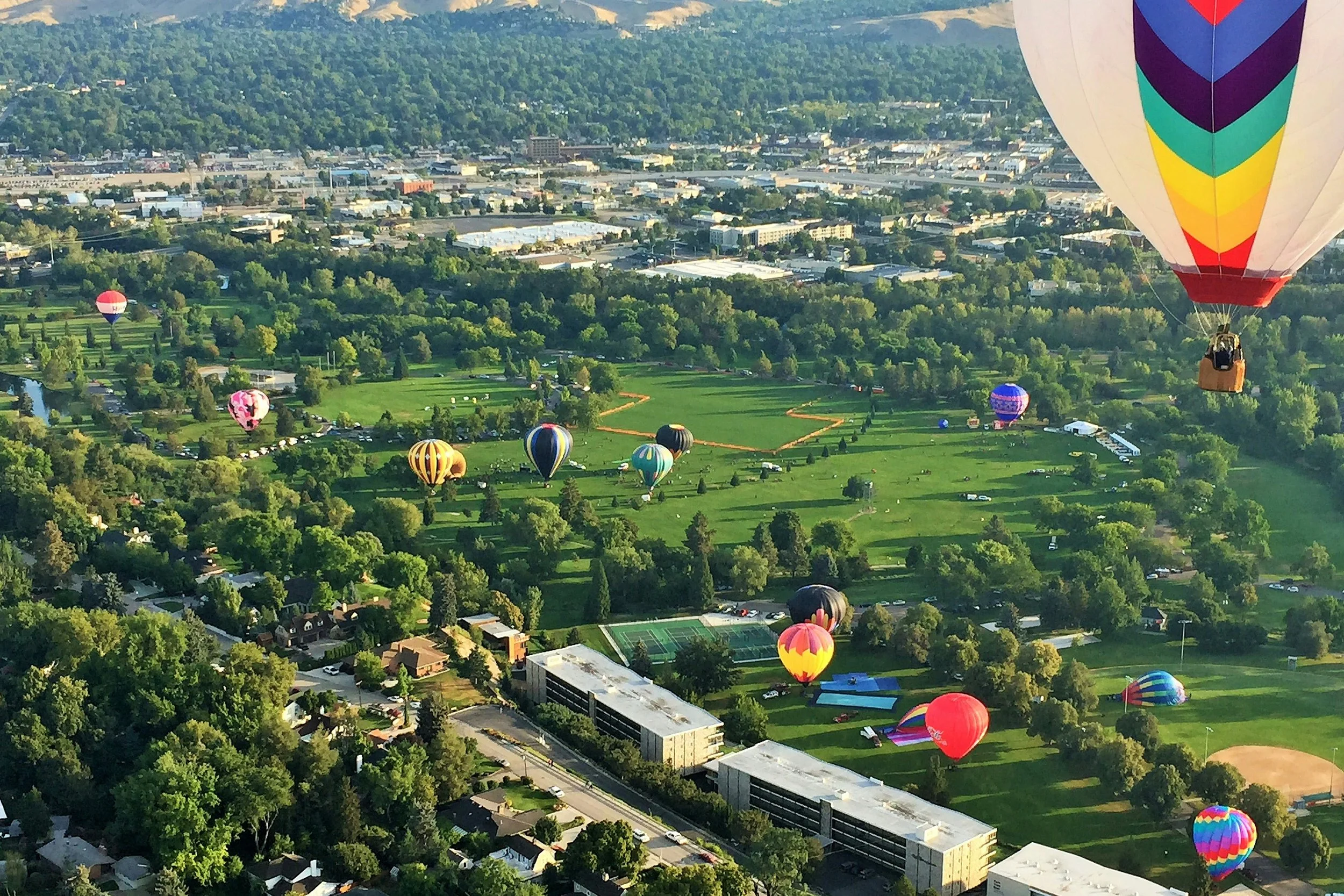 Boise, Idaho hot air balloons flying over Ann Morrsion Park with trees, tennis courts, and surrounding buildings and streets.