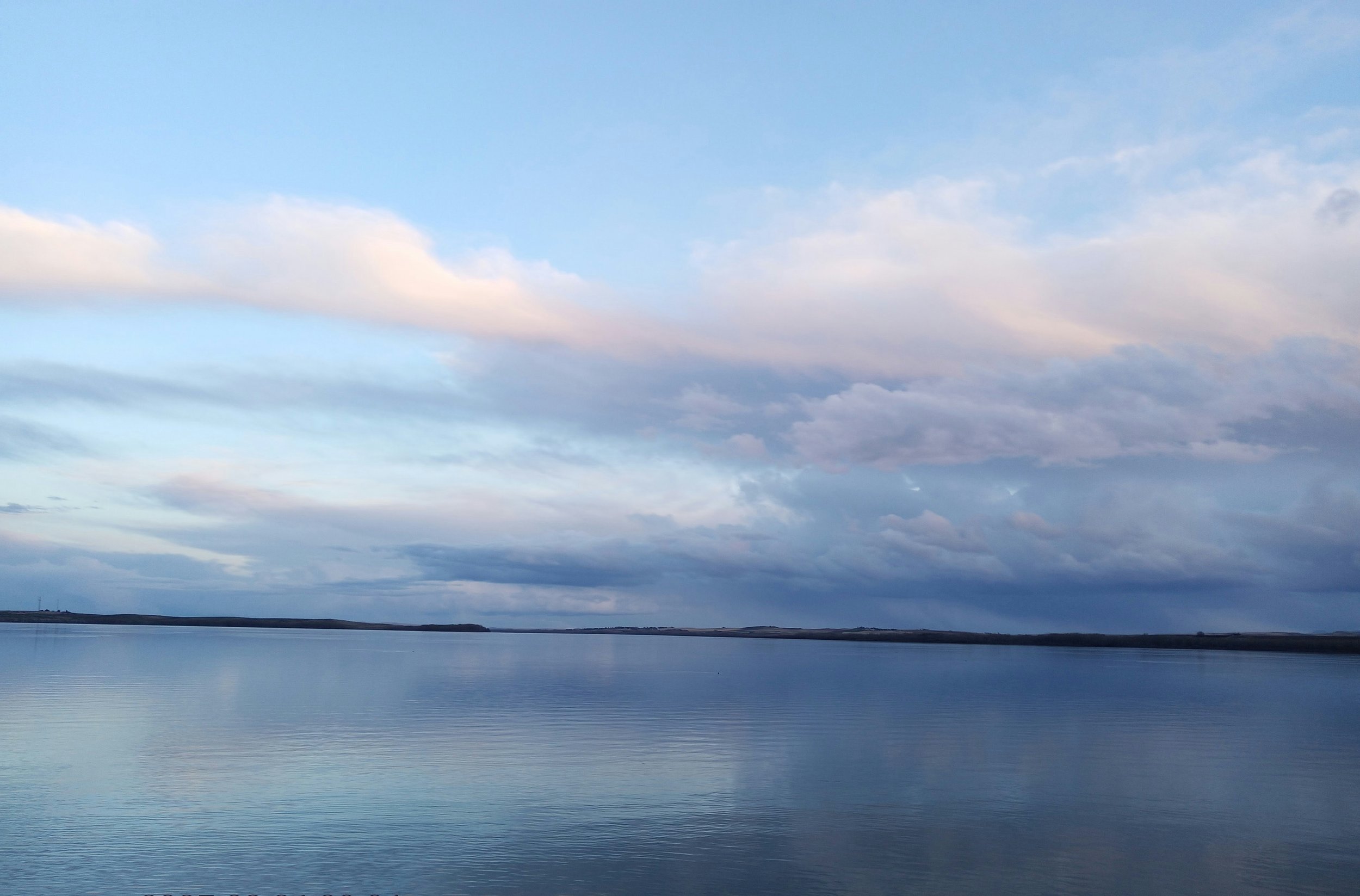 A calm body of water reflecting a partly cloudy sky with shades of blue and white, distant shoreline on the horizon.