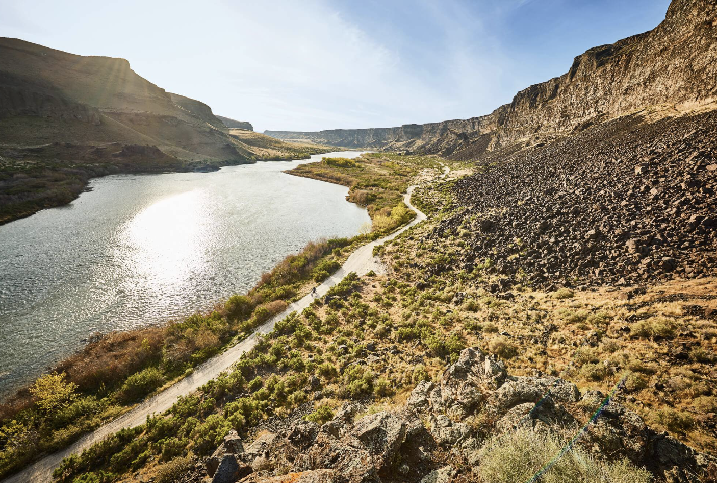 Scenic view of a river winding through a canyon with steep, rocky cliffs on both sides, some vegetation along the riverbank and a dirt trail beside the water under a clear blue sky.