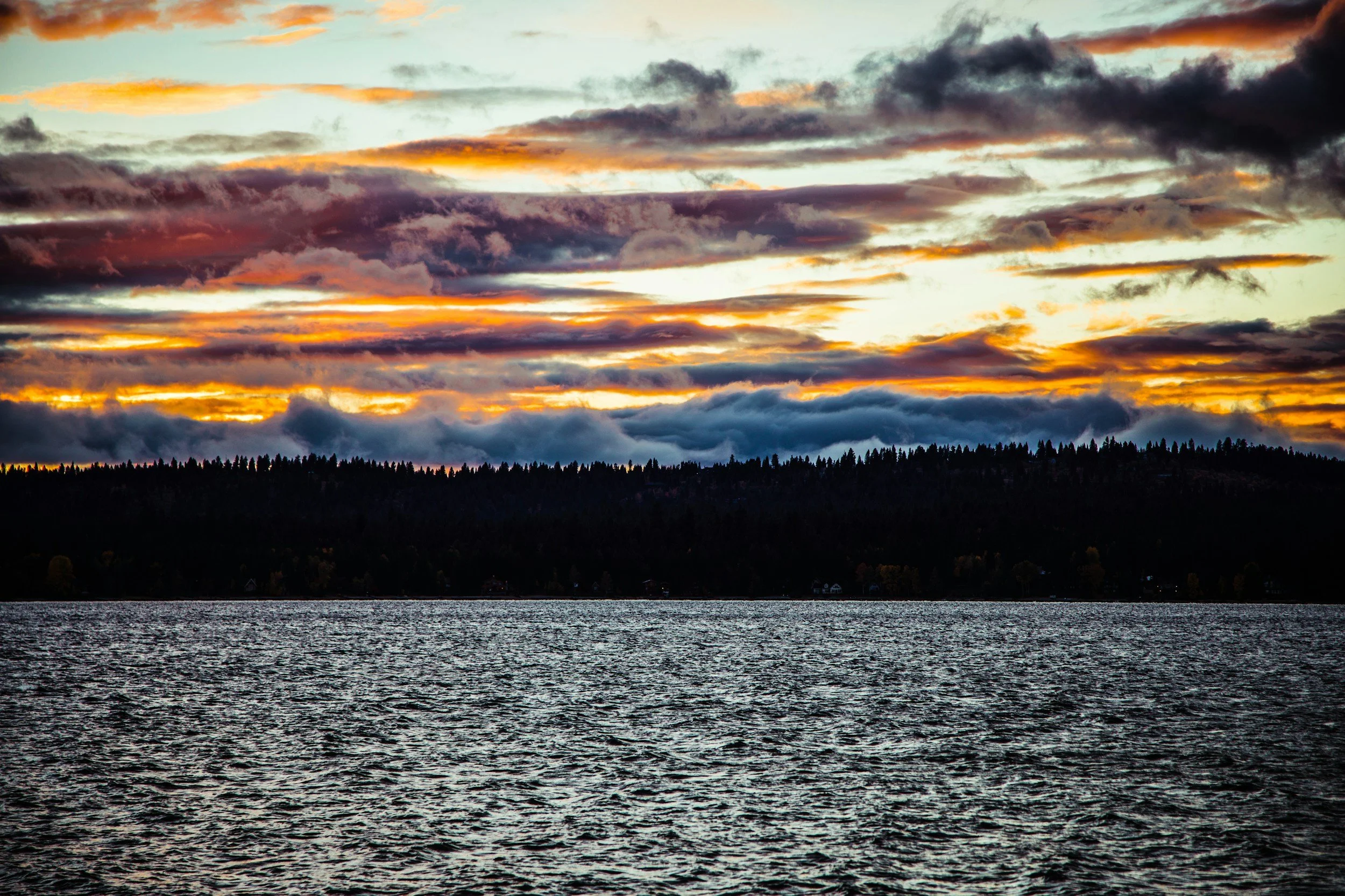 Sunset over a body of water with a silhouette of a forested shoreline and colorful clouds in the sky.