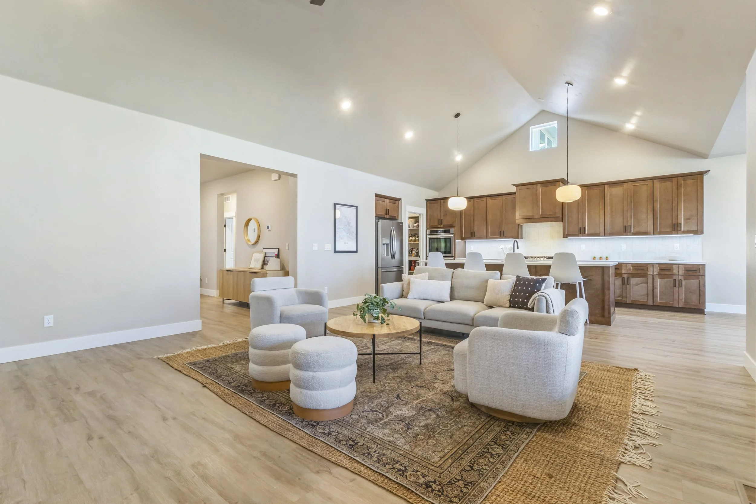 Living room and open kitchen with wooden cabinets, bar stools, and modern furniture including a sofa, armchairs, and ottomans, with hardwood flooring and a decorative rug.