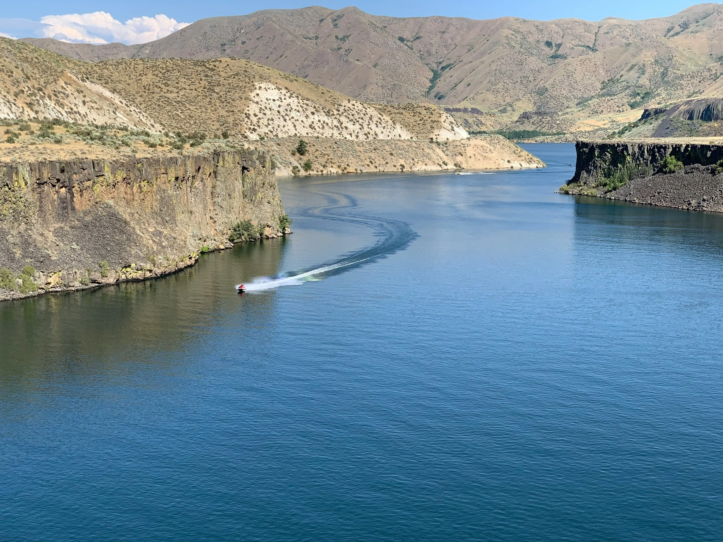 A river flowing through a canyon surrounded by dry, hilly terrain with mountains in the background and a boat creating a wake in the water.