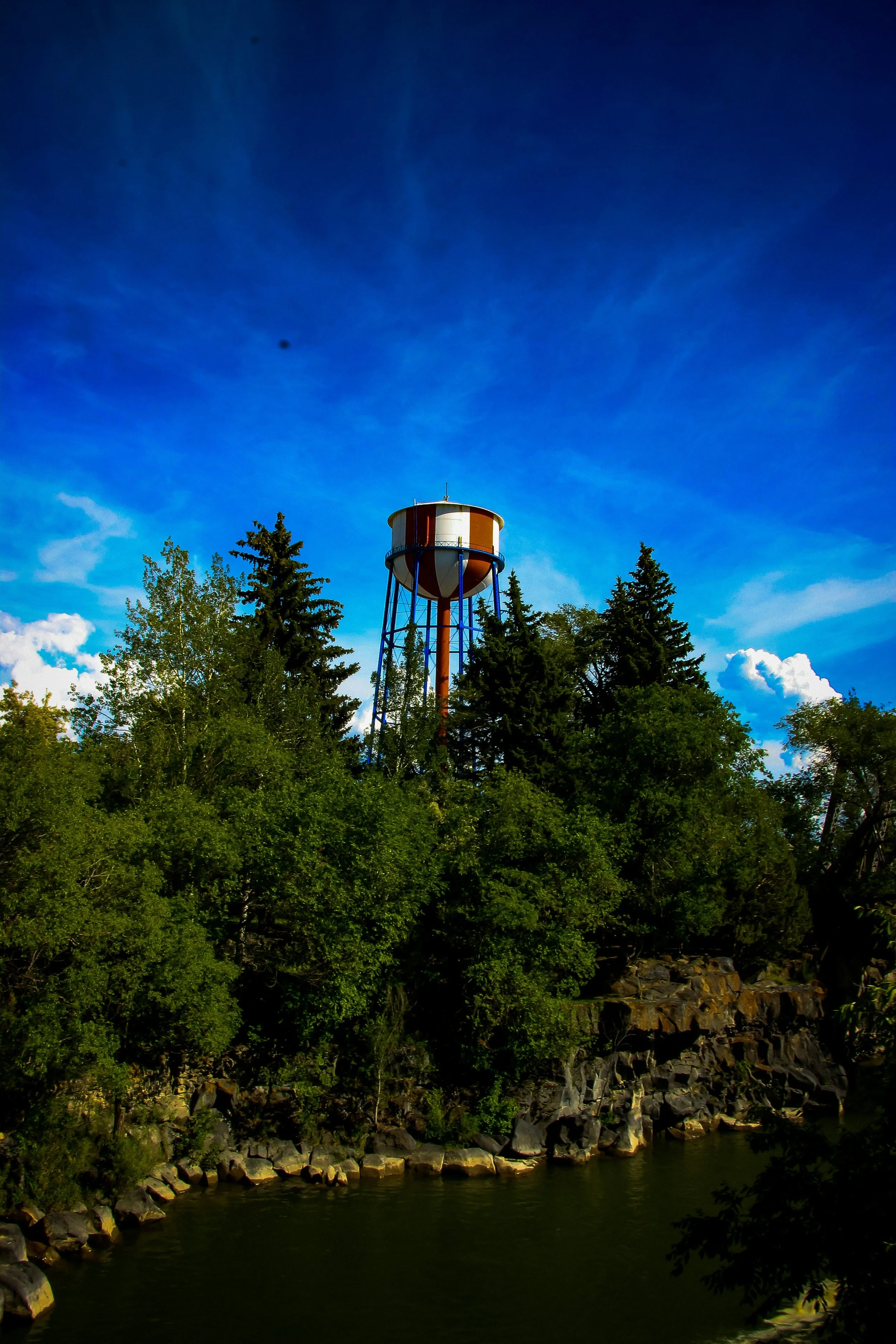 A water tower stands among tall green trees on a rocky shore next to a body of water under a bright blue sky with scattered clouds.