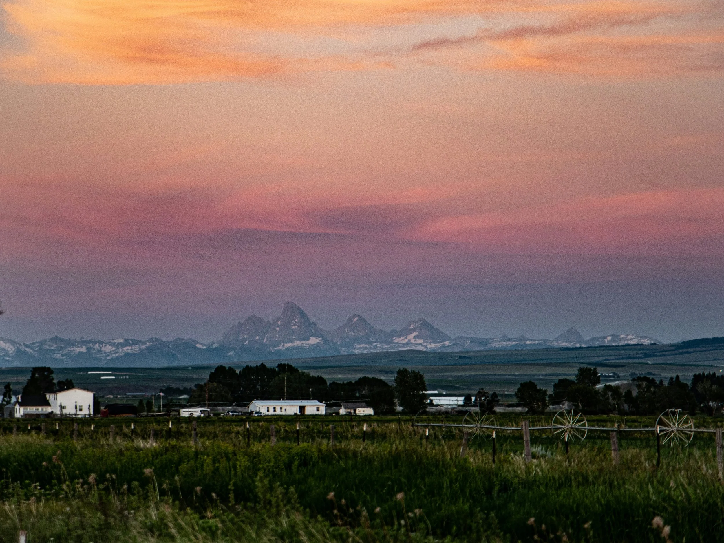 Sunset over open farmlands with purple mountains in the background, farm buildings, trees, and a wire fence in the foreground.