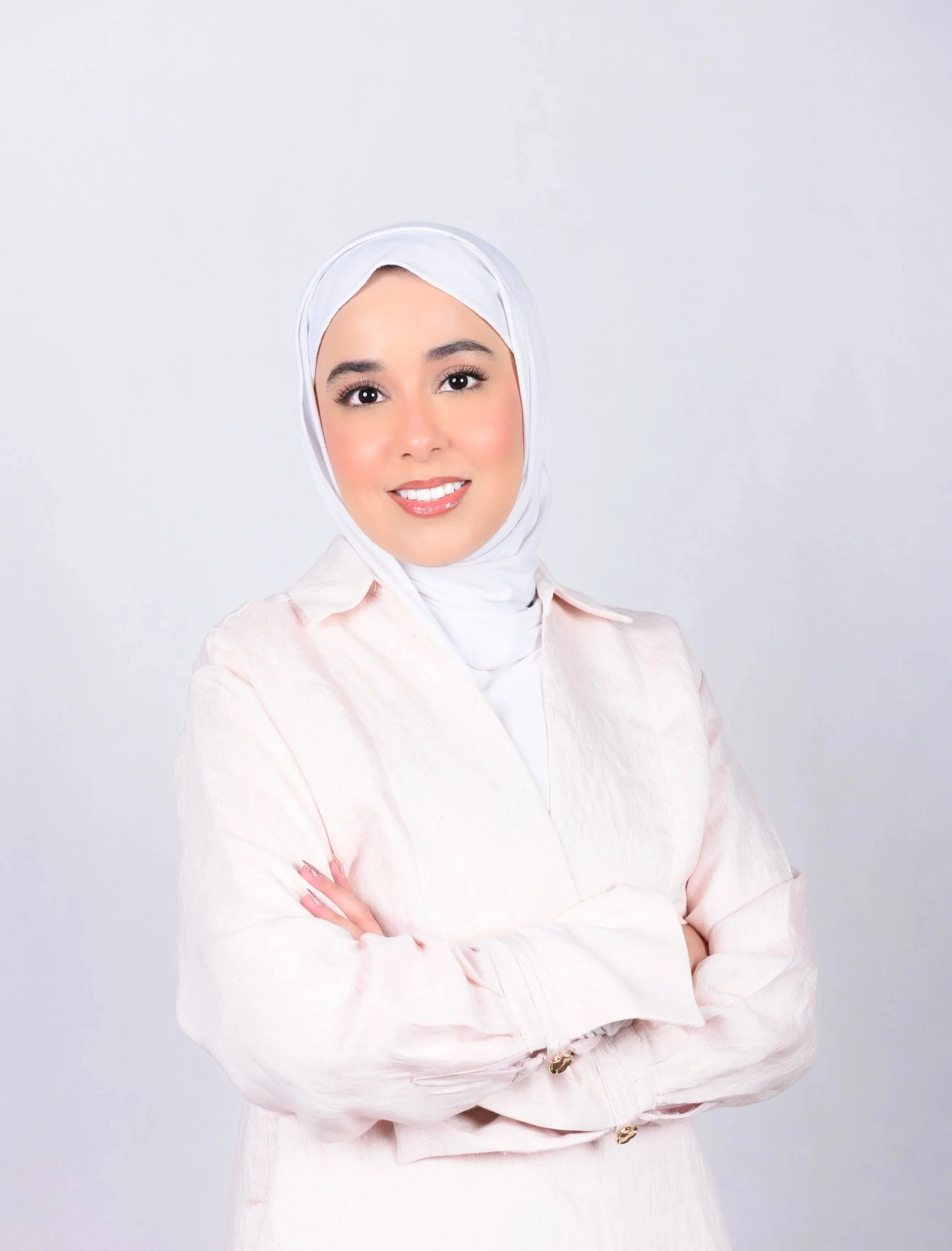 A woman wearing a white hijab and light pink jacket, smiling with arms crossed against a plain light background.