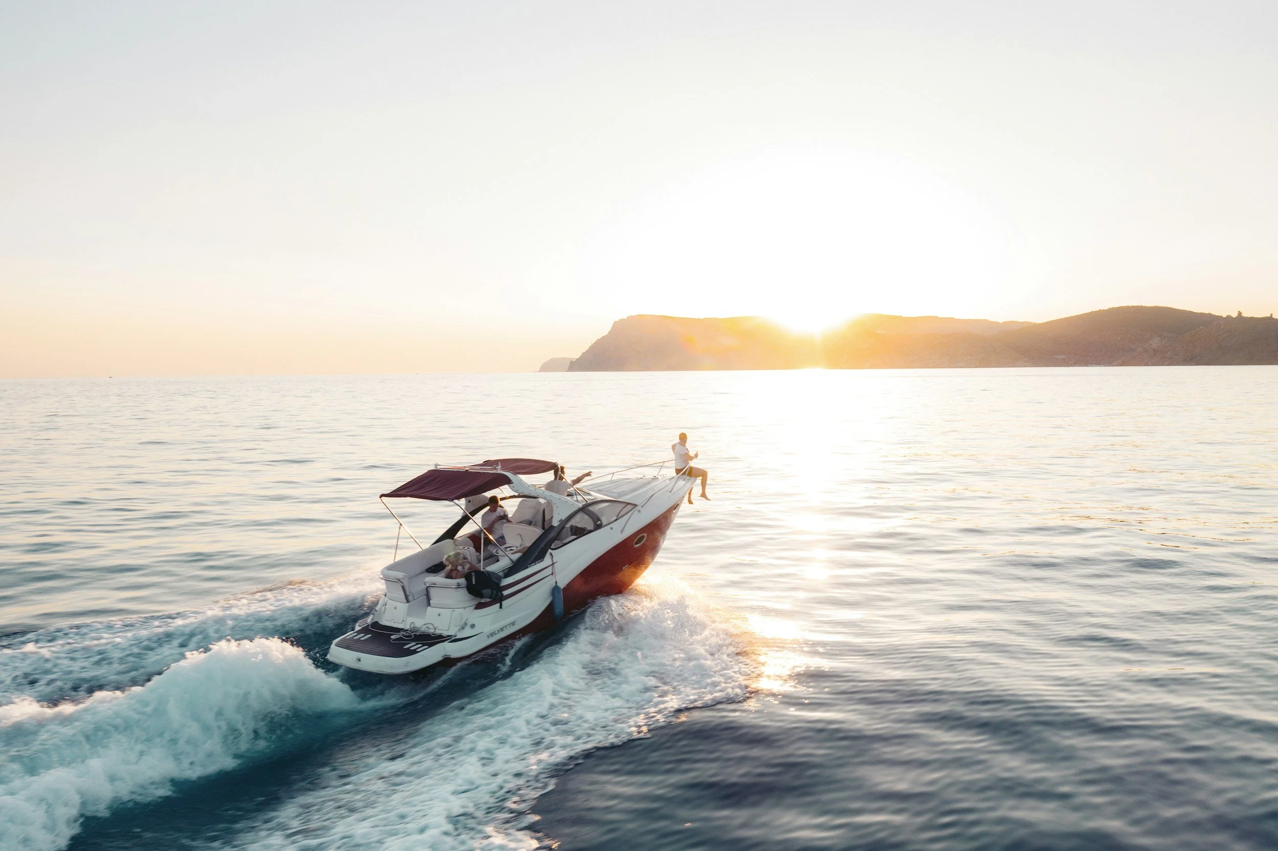 A motorboat cruising on the ocean at sunset with a person sitting on the bow and another in the boat, and a distant island silhouette in the background.