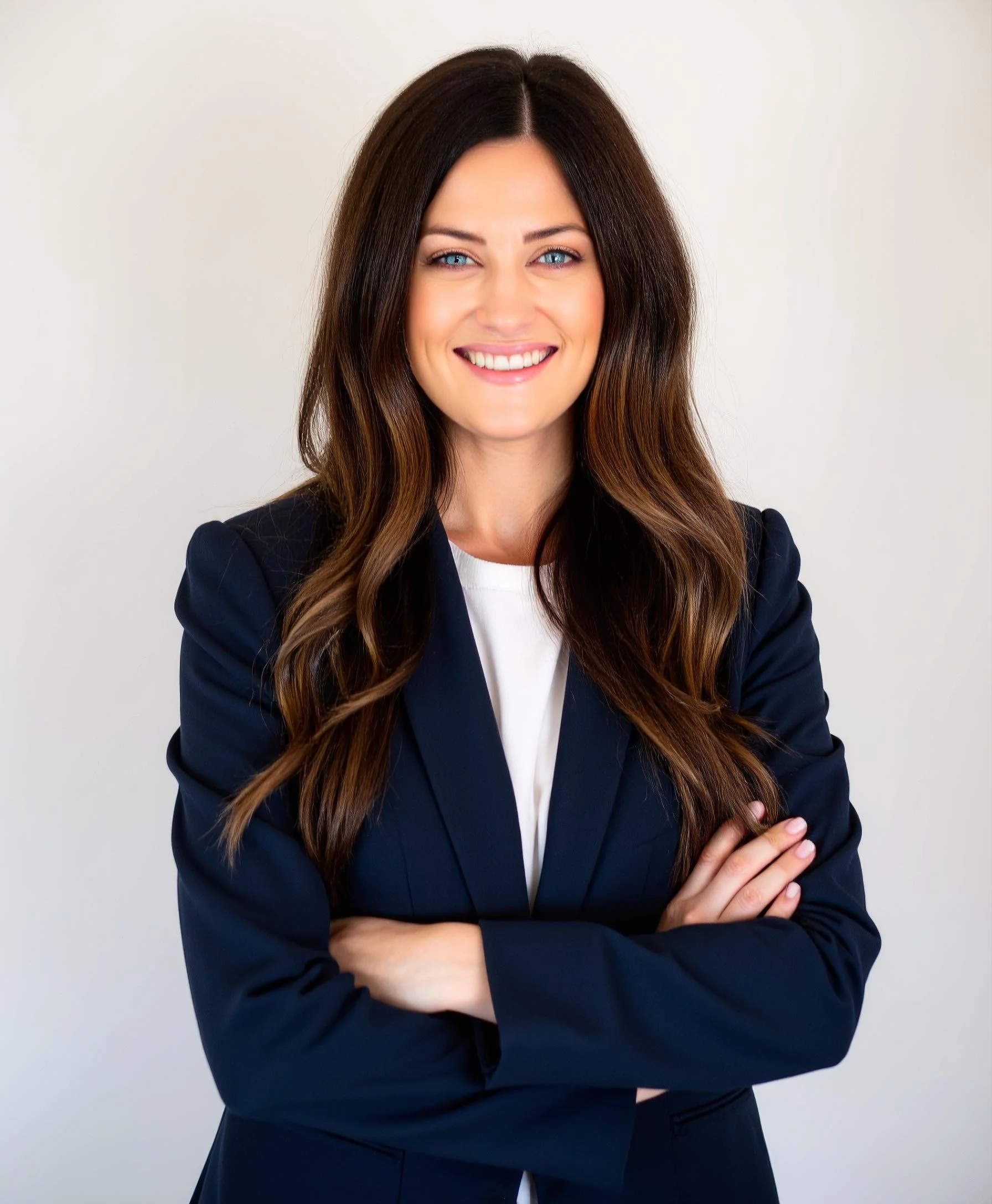 A confident young woman with long brown hair, wearing a navy blazer and a white top, smiling with her arms crossed against a plain white background.