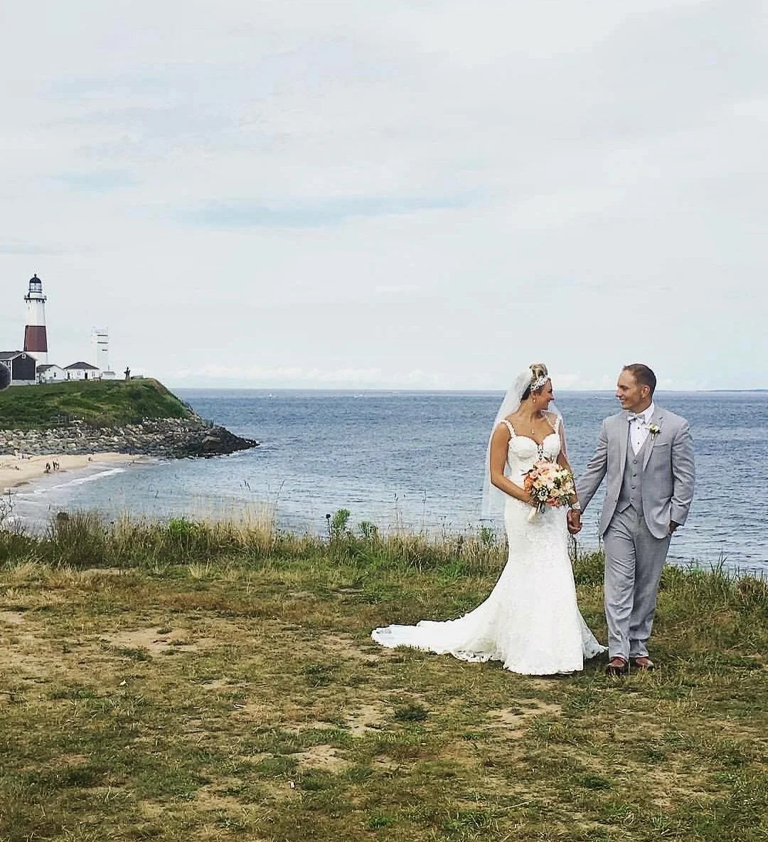 A bride and groom holding hands by the seaside with a lighthouse in the background, on a cloudy day.