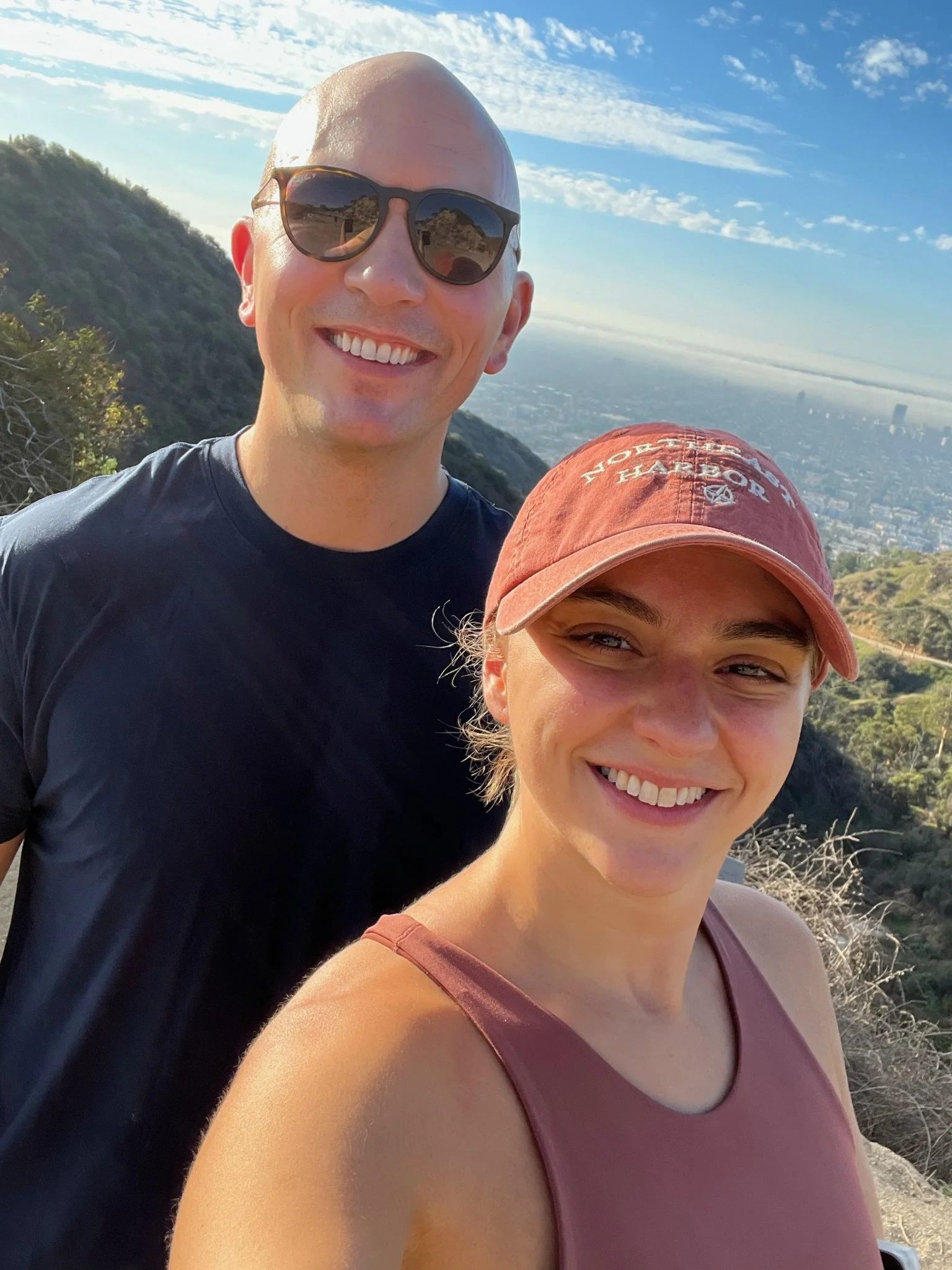 Two smiling young adults, a man and a woman, taking a selfie on a mountain trail with a city skyline and blue sky in the background.