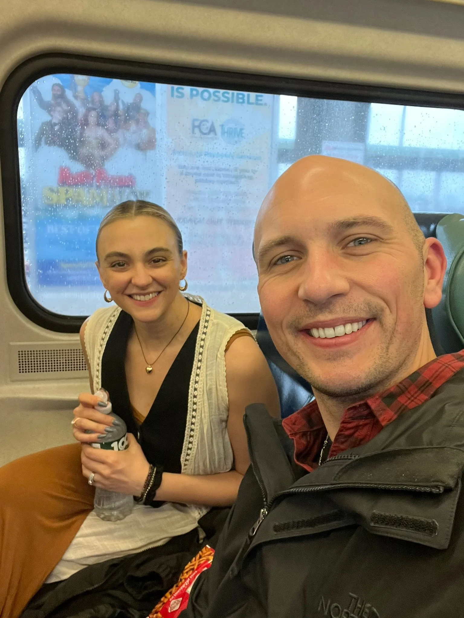 A smiling man and woman taking a selfie on a train, with a rainy window behind them. The woman holds a water bottle and wears a sleeveless top with a cardigan, and the man wears a black jacket and red plaid shirt.