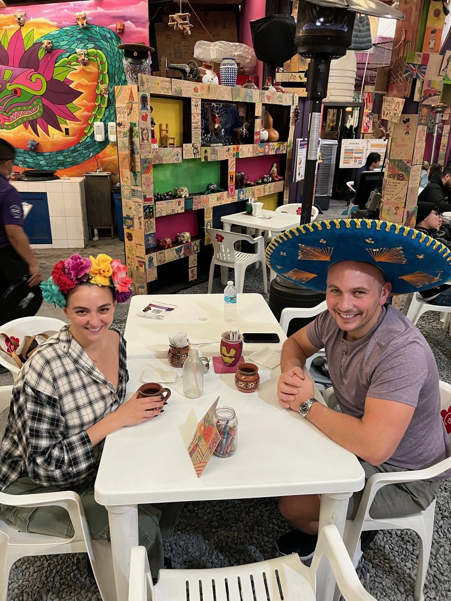 A man and woman sitting at a table in a colorful restaurant, both wearing Mexican-themed hats, smiling at the camera. The woman has a floral headband, and the man has a large blue sombrero. The table has drinks, decoration, and a small card.