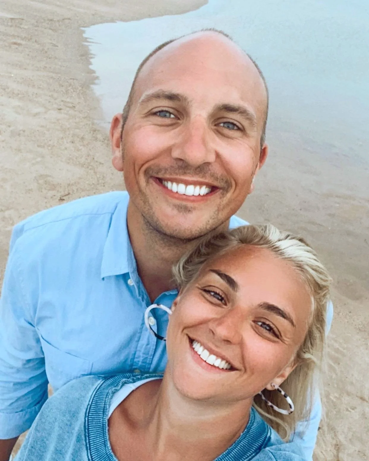 A smiling man and woman taking a selfie at the beach, with the shoreline and water in the background.