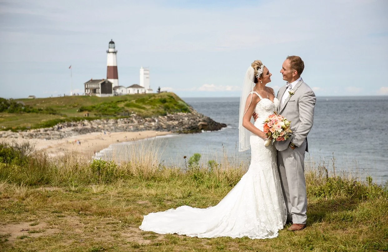 A bride and groom standing together on a grassy area near the coast, holding a bouquet, with a lighthouse and ocean in the background.