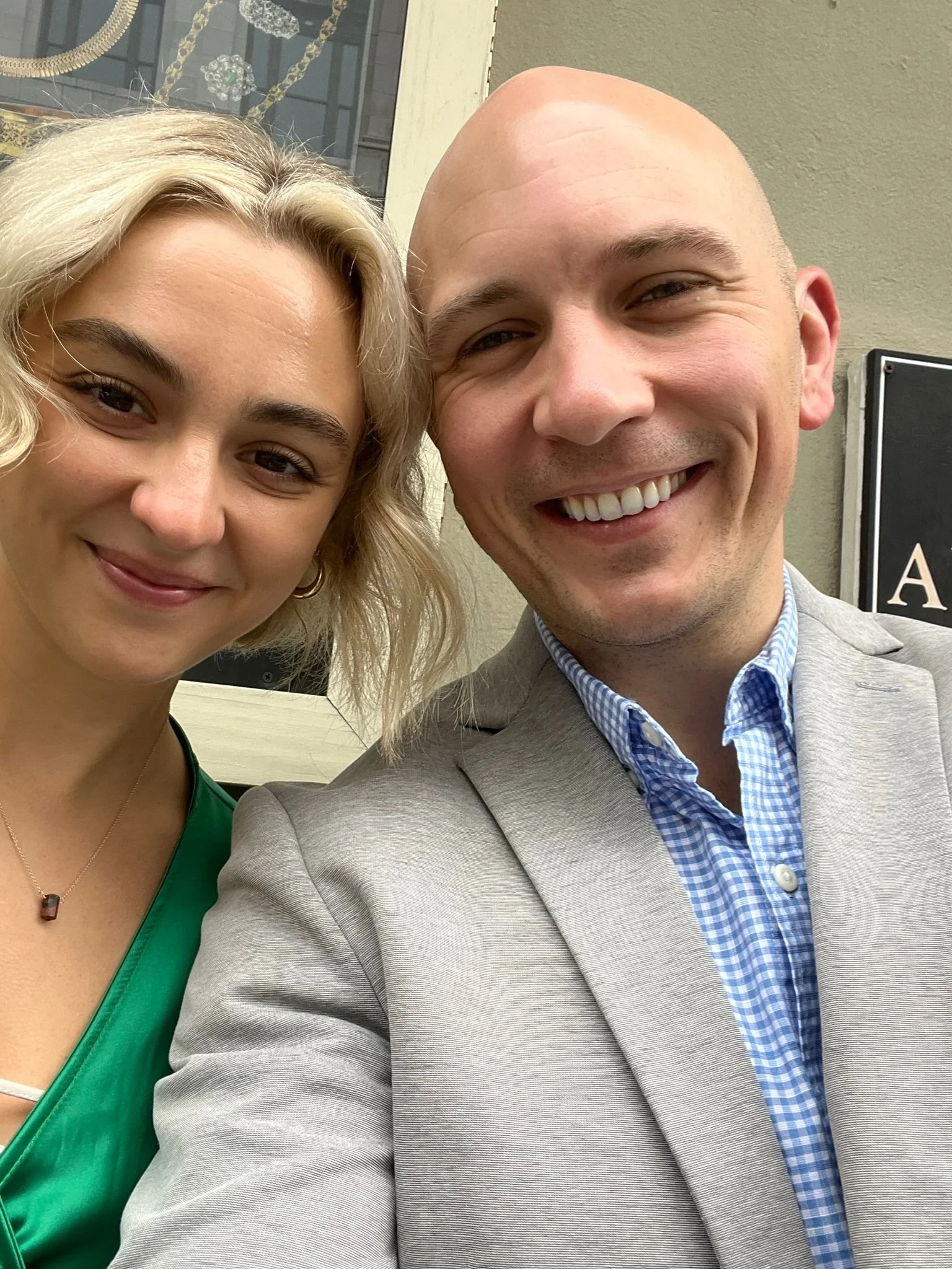 A smiling man and woman taking a selfie outdoors with buildings in the background.