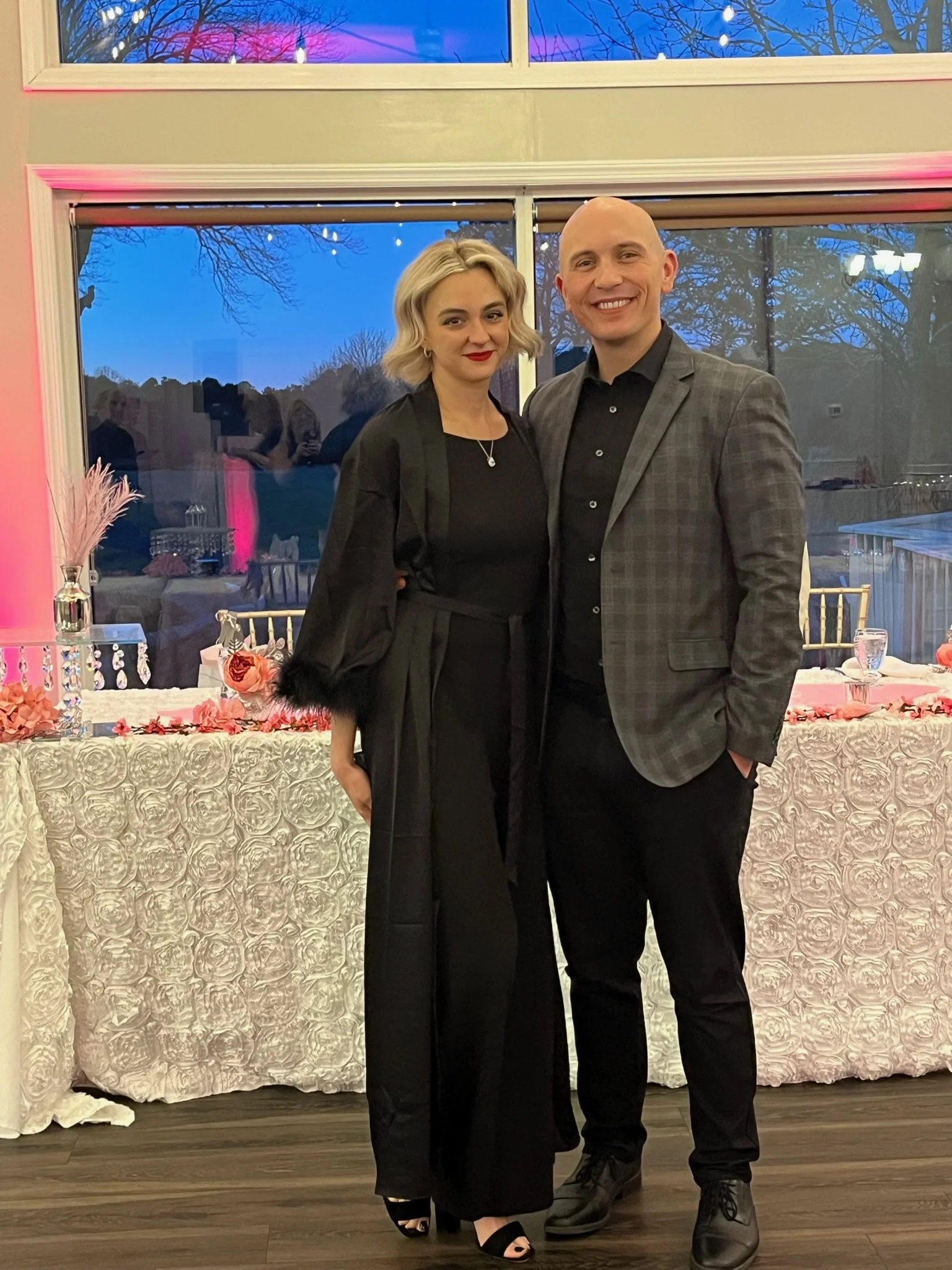 A man and woman standing together in formal attire, smiling at the camera, at an indoor event with decorated tables and a large window showing evening sky and trees outside.