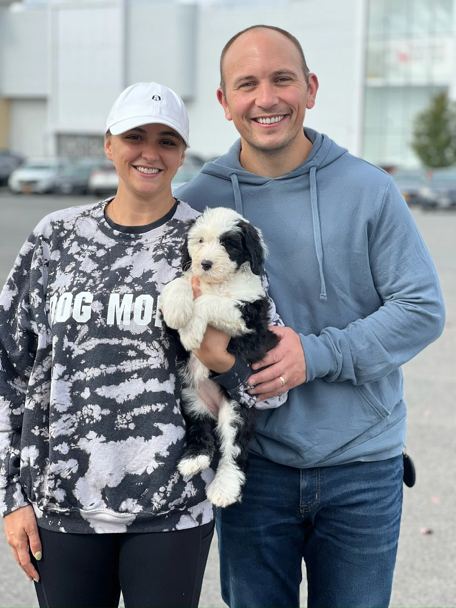 A smiling woman wearing a white cap and black and white tie-dye sweatshirt holding a black and white puppy, standing next to a smiling man in a blue hoodie in an outdoor parking lot.