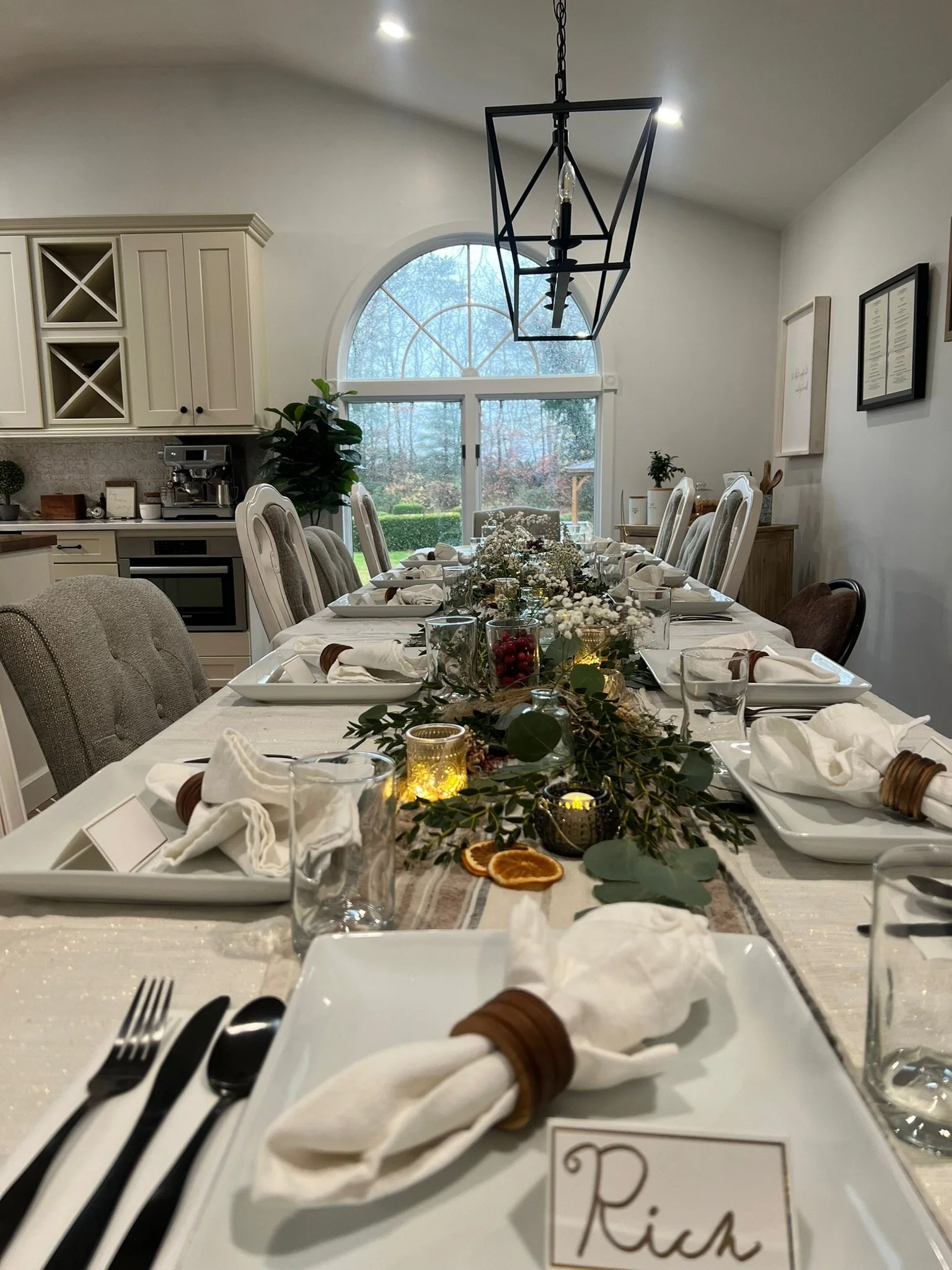 Dining table set for a formal gathering with white plates, cloth napkins with wood ring napkin holders, glasses, and a greenery and candle centerpiece, in a bright room with large window and modern chandelier.