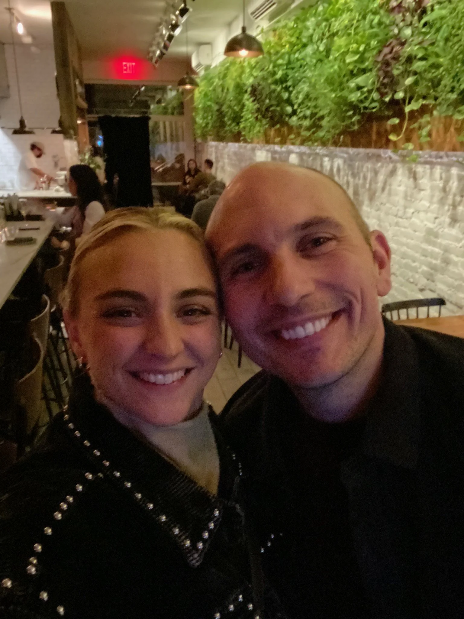 A smiling woman and man take a selfie in a dimly lit restaurant with white brick walls and green plants.