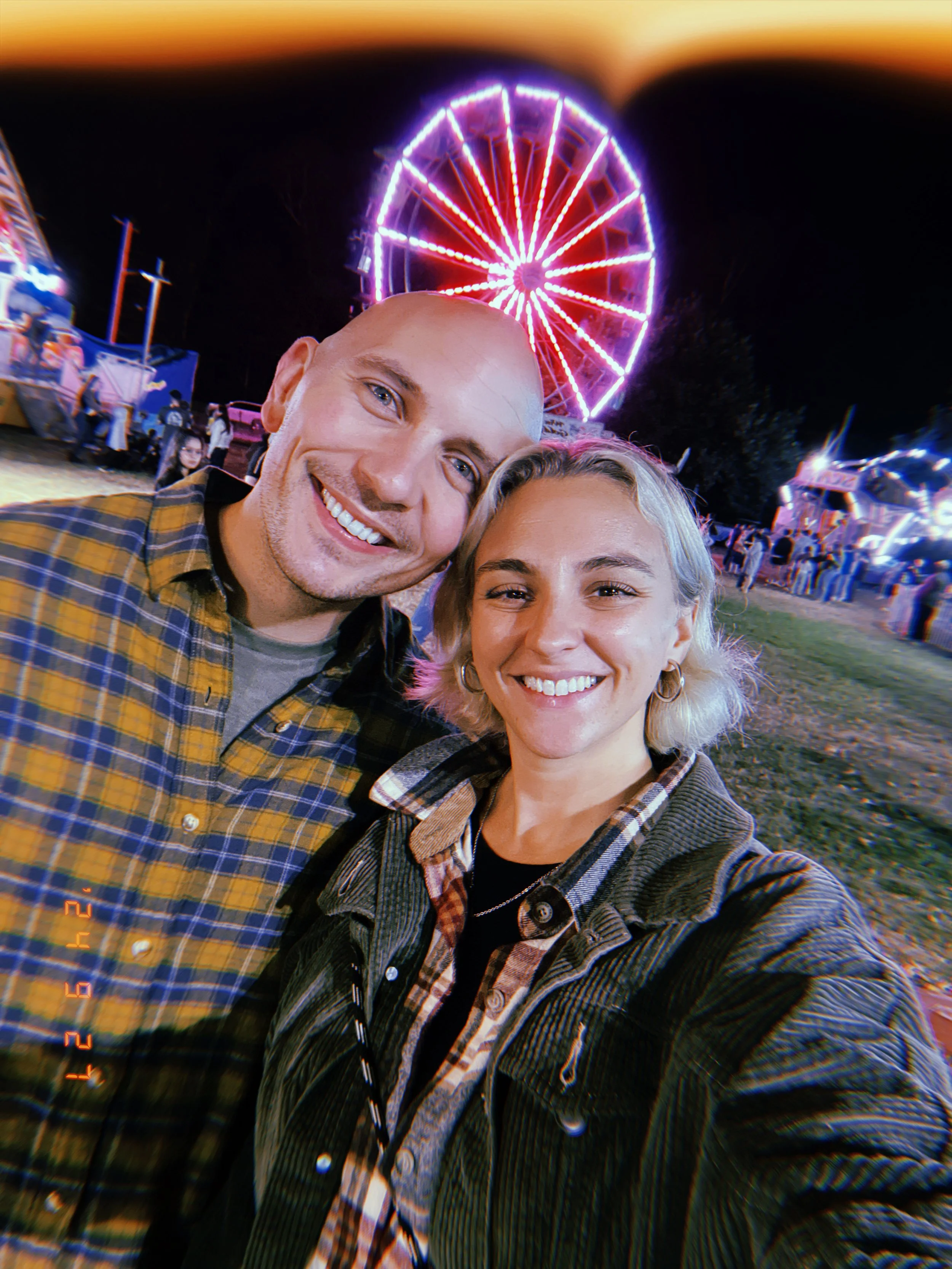 A smiling couple taking a selfie at a carnival at night, with a brightly lit Ferris wheel in the background.