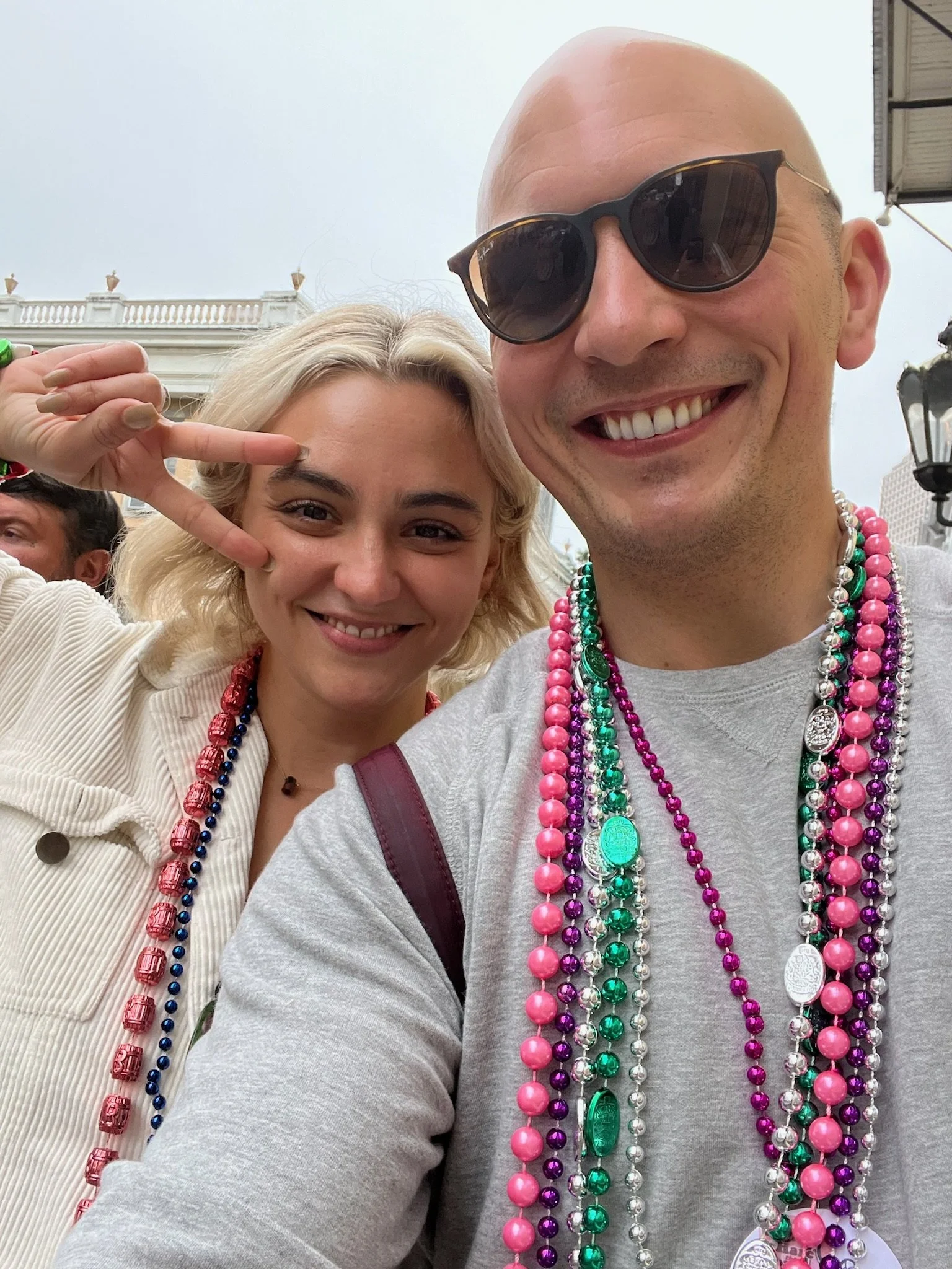 Two smiling people taking a selfie together, wearing colorful Mardi Gras bead necklaces, with a city building in the background during daylight.