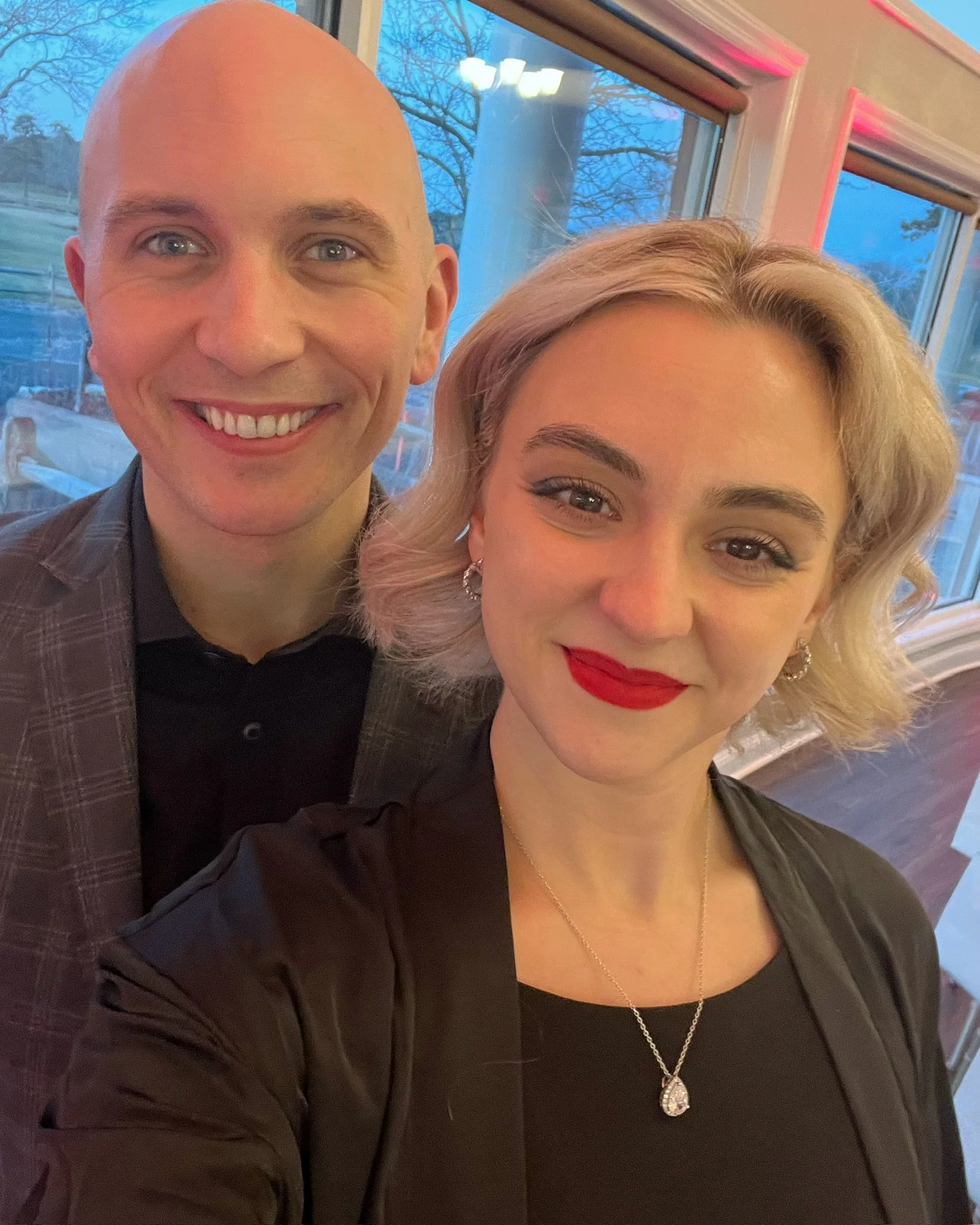 A man and woman taking a selfie indoors, with large windows showing a blue sky and trees outside.