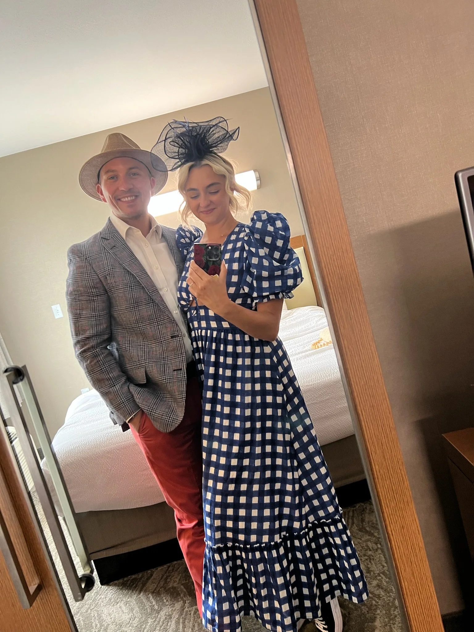 A man and woman taking a selfie in a hotel room mirror, both dressed in fashionable attire with hats. The man wears a gray checked blazer and a straw hat, while the woman wears a blue and white checkered dress with puffy sleeves and a black fascinato