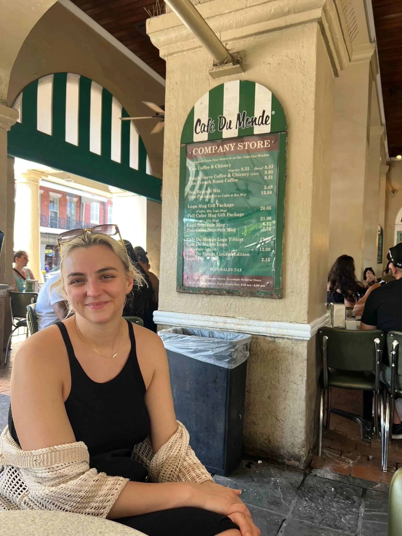 A woman sitting at an outdoor café table, smiling, with a menu on the wall behind her and other people dining nearby.