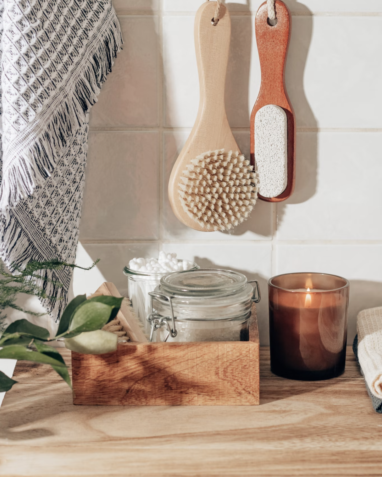 Bathroom countertop with hanging brushes, a candle, a glass jar, and a tray with cotton balls, with a towel and green plant leaves visible.