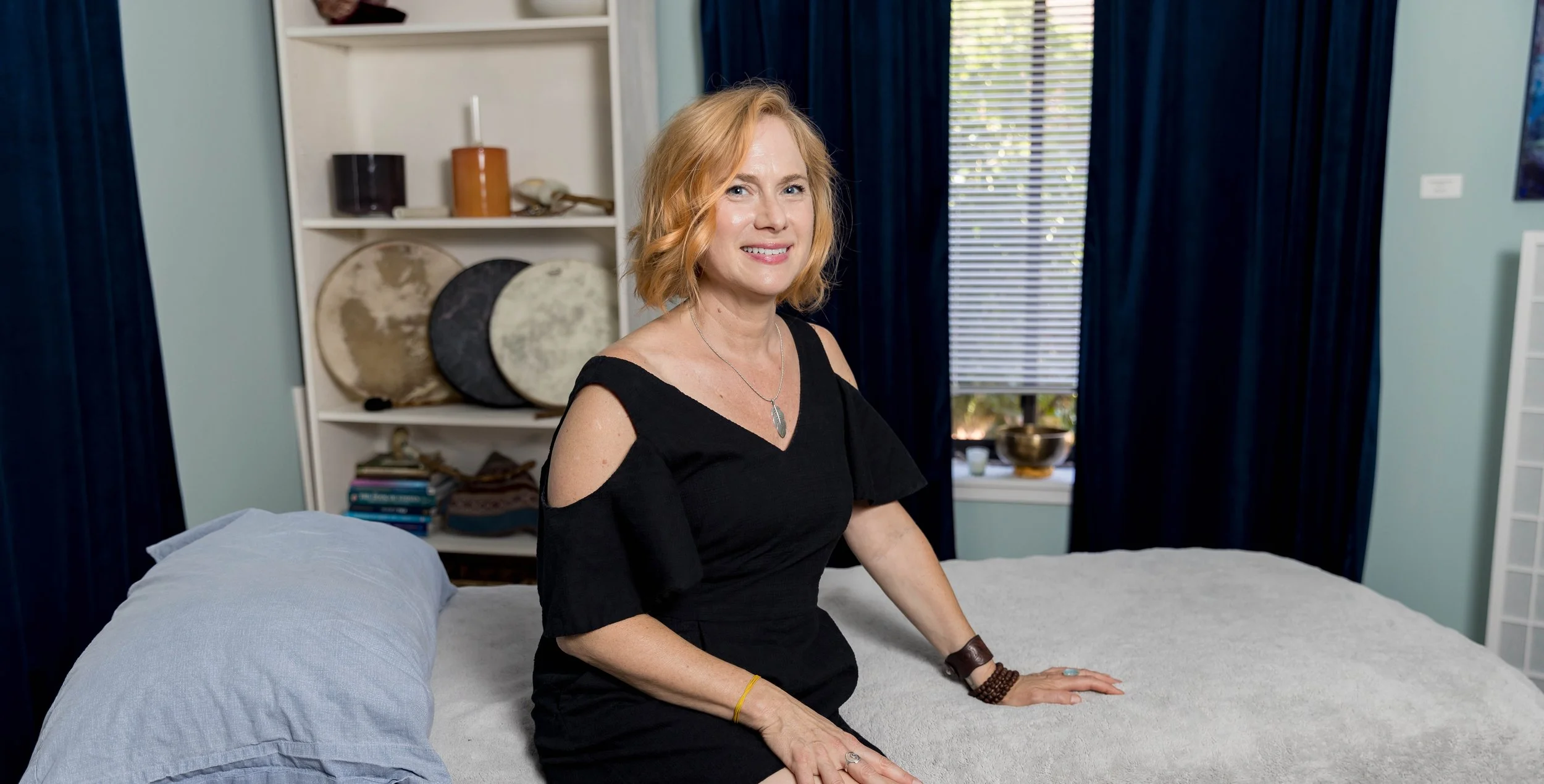 A woman with shoulder-length blonde hair, wearing a black dress with cut-out shoulders, sitting on a bed in a bedroom. She is smiling and looking at the camera. There are blue curtains, a window with blinds, and decorative items on a shelf in the background.