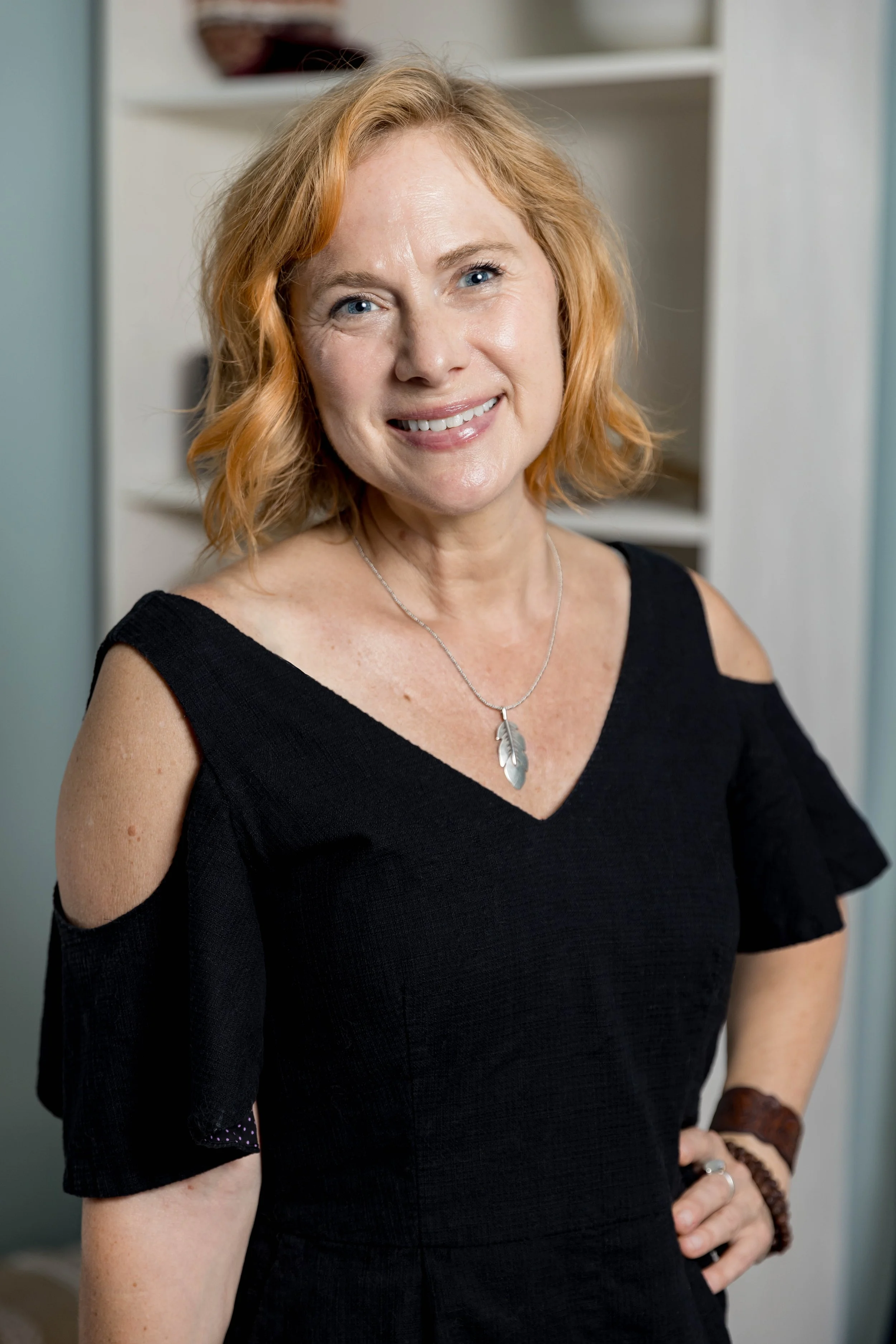 A woman with short, curly red hair smiling at the camera, wearing a black cold-shoulder top with a necklace featuring two feathers, standing in a room with shelves in the background.
