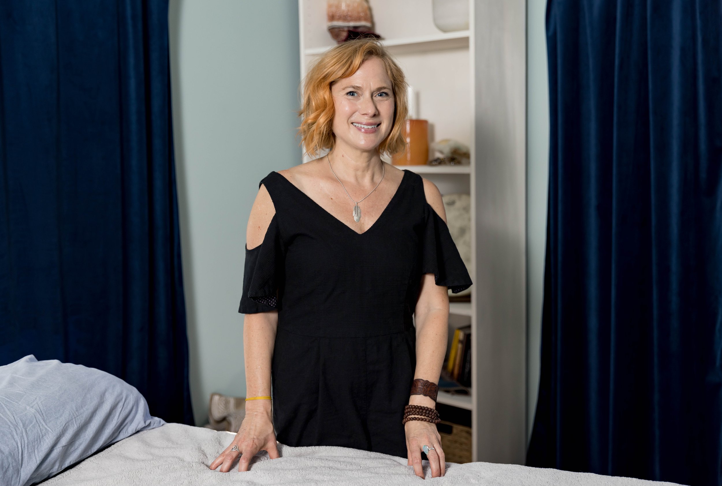 A woman with shoulder-length red hair, wearing a black dress with cut-out shoulders, stands in a bedroom, smiling with her hands resting on a bed, with dark blue curtains and a bookshelf in the background.