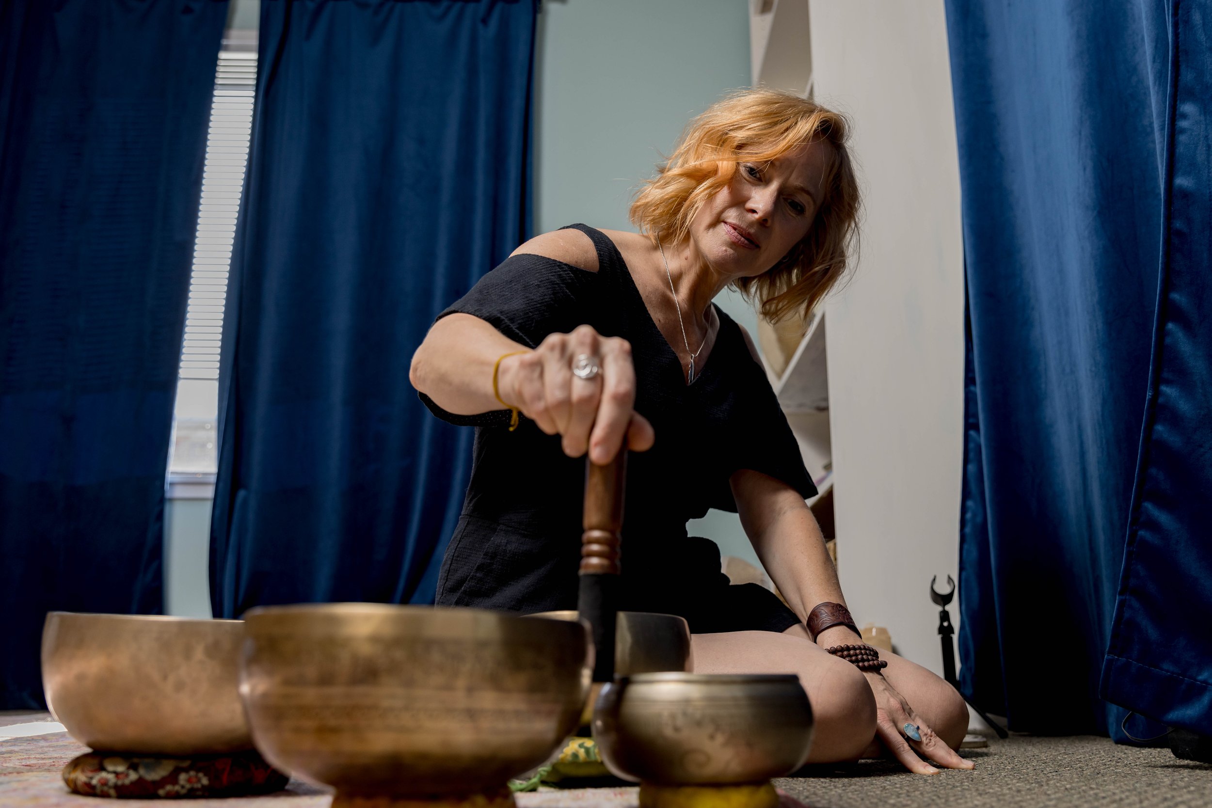 A woman with red hair and wearing a black dress is sitting on the floor playing singing bowls.