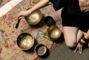 Person sitting on a rug with six singing bowls of different sizes and colors, holding a mallet.