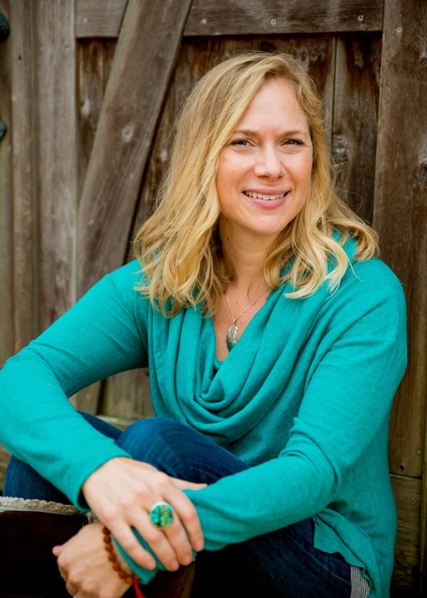 A woman with blonde hair smiling, sitting outdoors against a wooden fence, wearing a teal long-sleeve top, jeans, and accessories including a large ring, a necklace, and a bracelet.