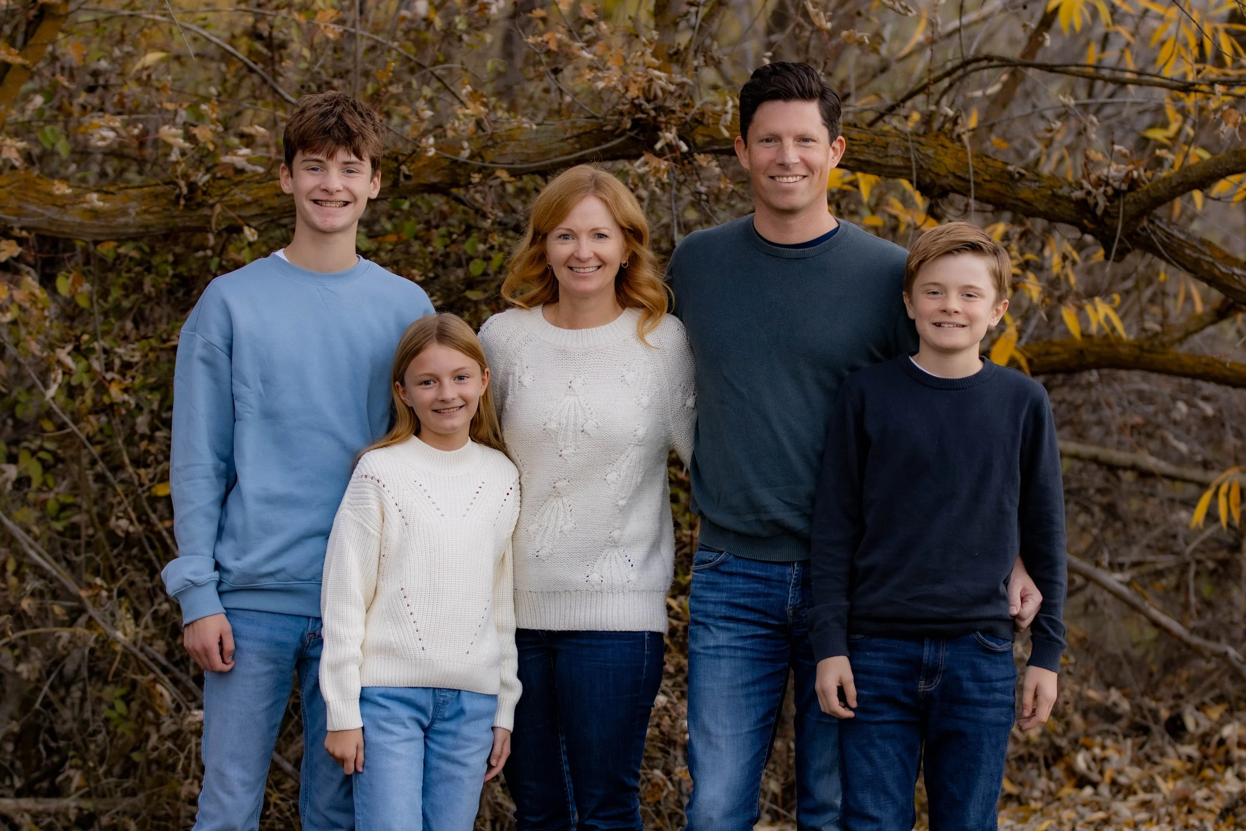 Family of five standing outdoors in fall, smiling, with trees and fallen leaves in the background.