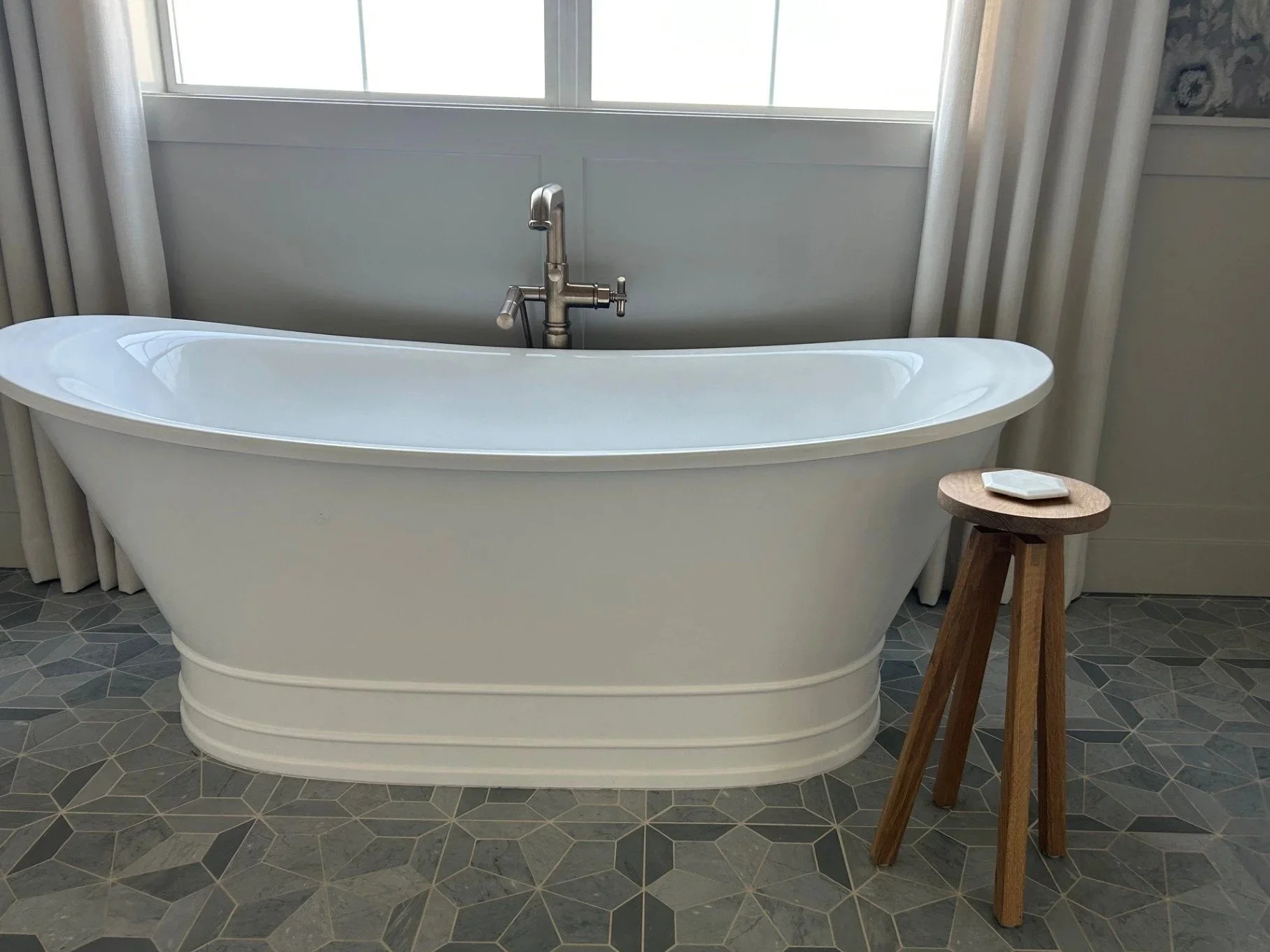 White freestanding bathtub with silver faucet and hand shower, positioned in a room with patterned tile flooring, curtains, and a wooden stool with a white dish on top.