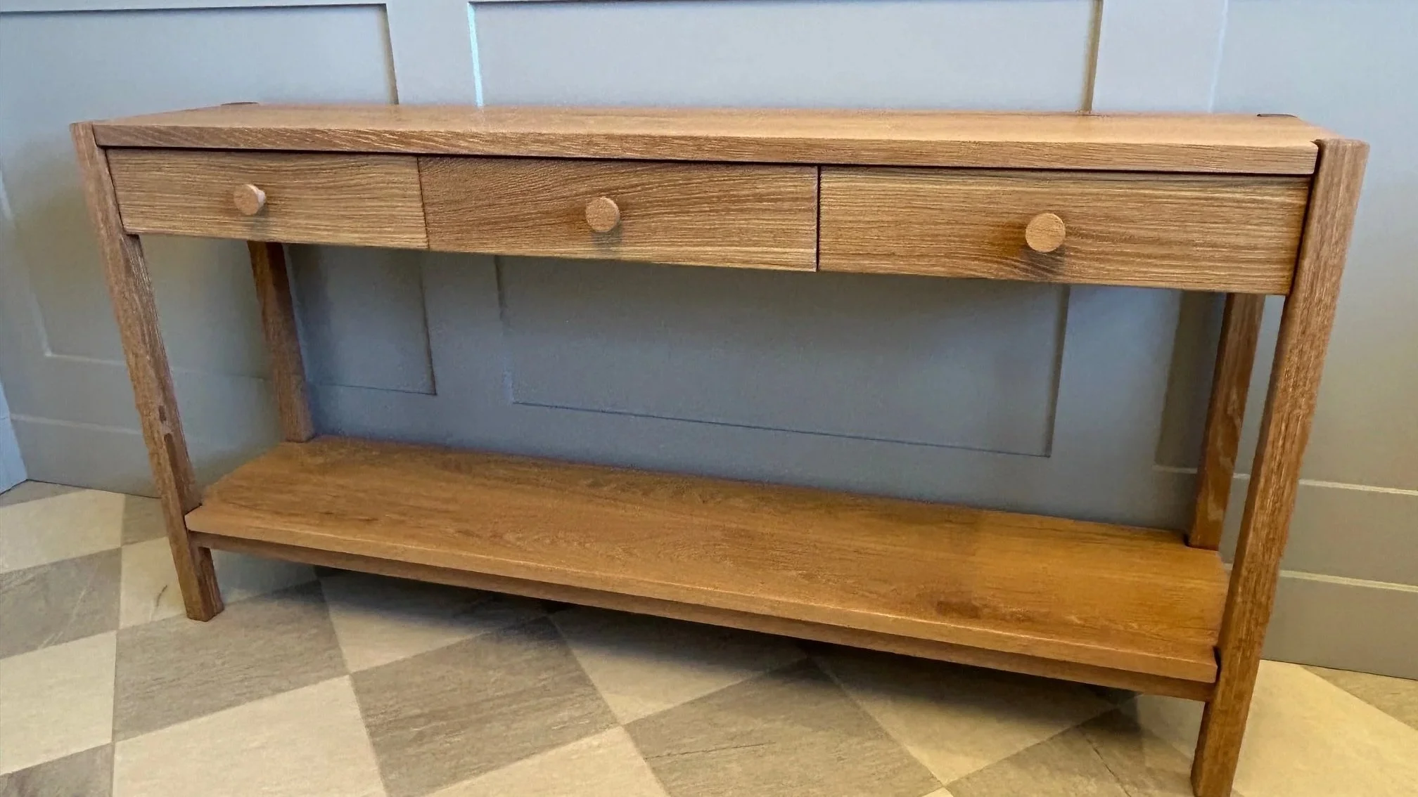 A wooden console table with three small drawers on top and a lower shelf, placed against a light gray wall and on a checkered tile floor.