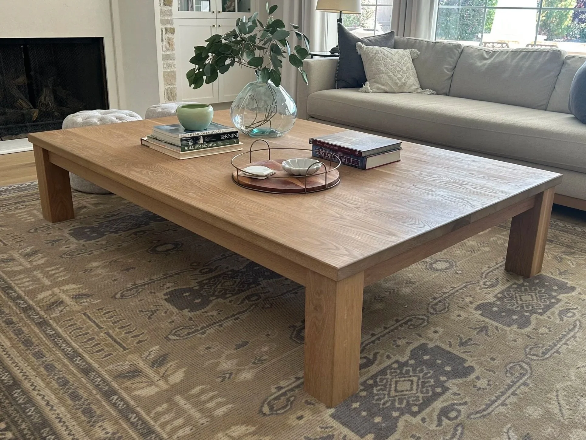 A living room with a large wooden coffee table on a beige patterned rug, a gray couch with pillows, a glass vase with greenery, and hardcover books on the table, with a fireplace in the background and large windows letting in natural light.