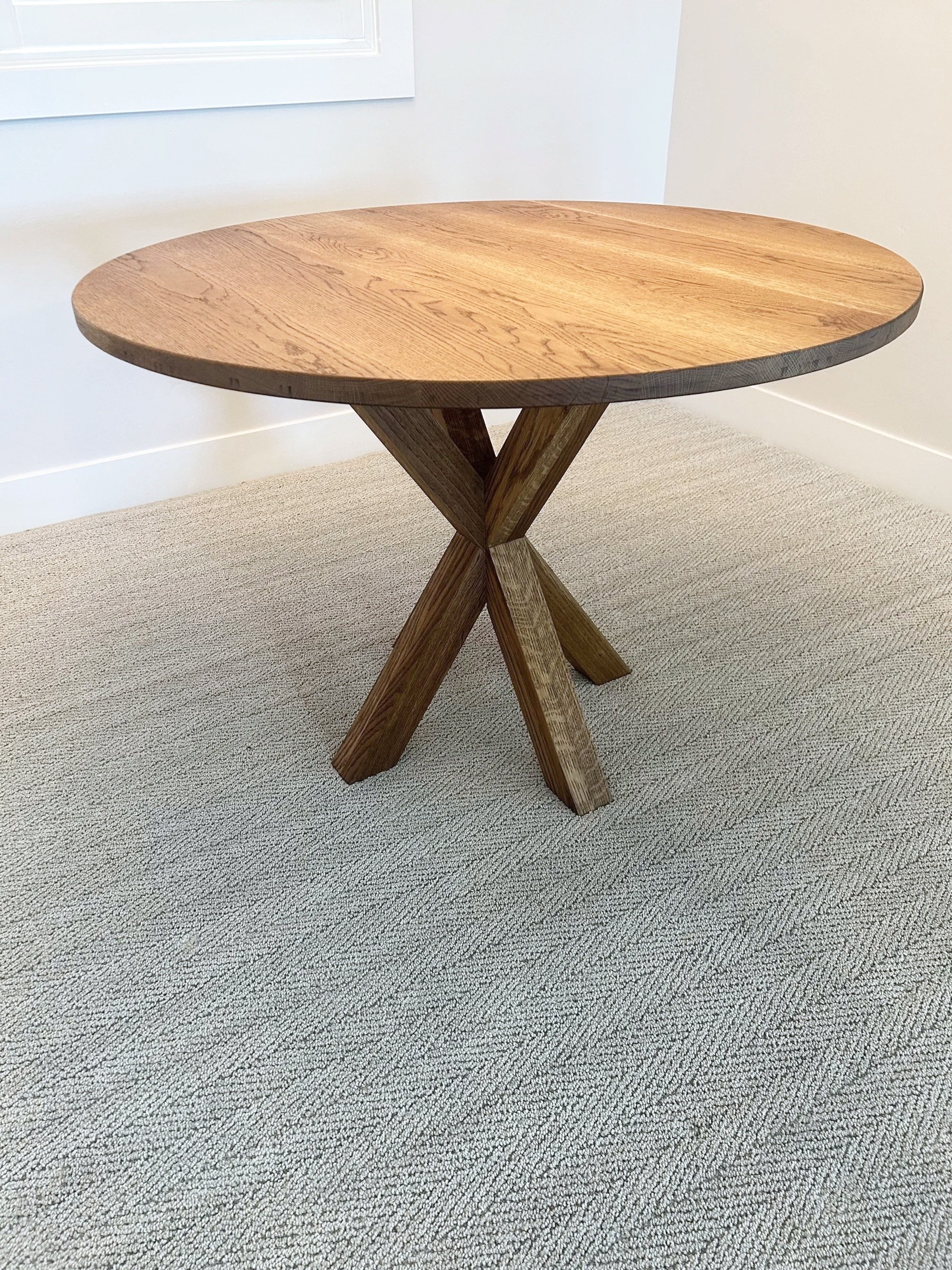 A round wooden table with a cross-leg wooden base, placed on a beige carpeted floor near a white wall.