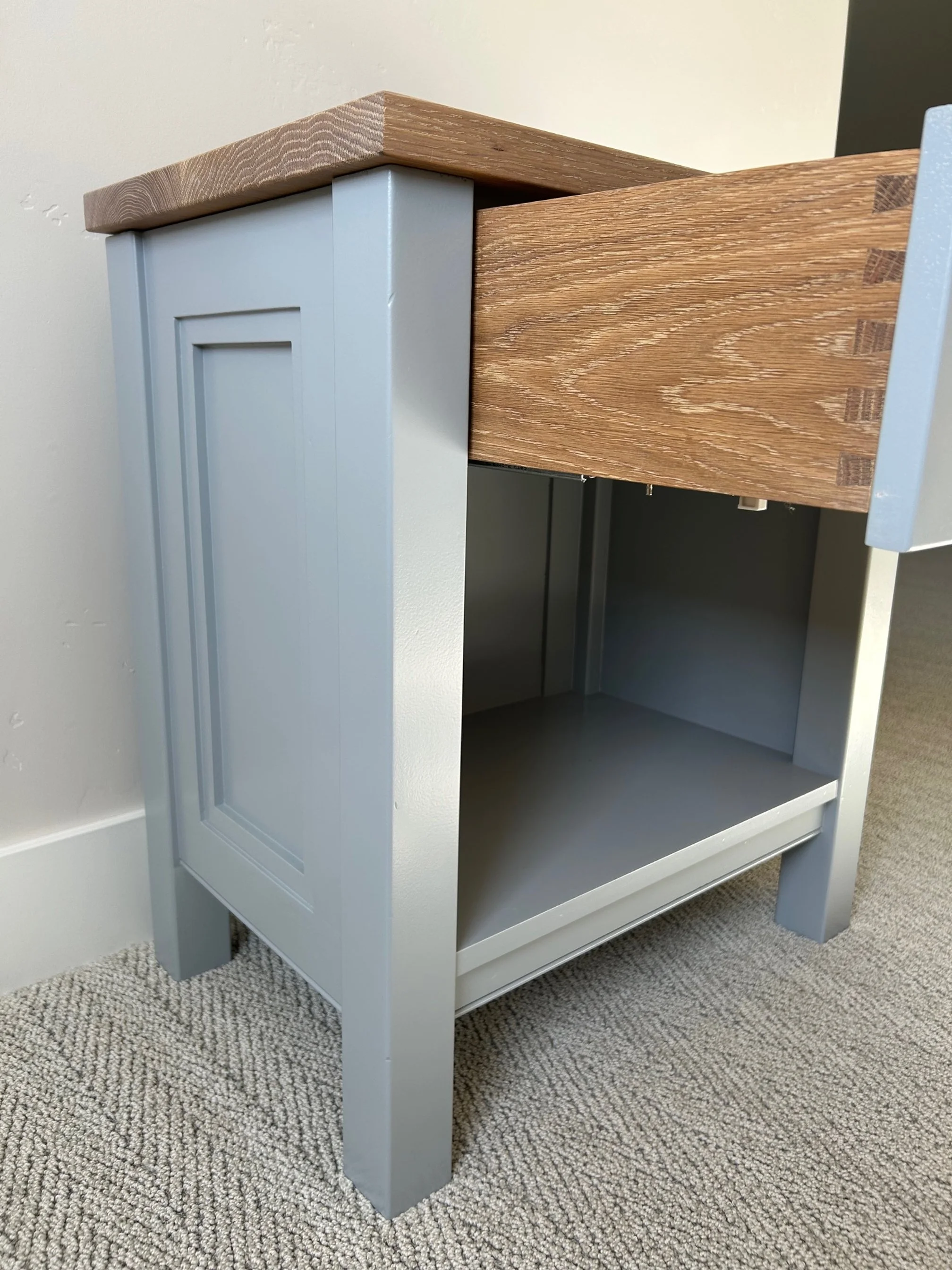 Close-up of a painted wooden cabinet with one door open, showing the interior. The cabinet has a blue-gray color and is placed on a beige carpeted floor against a white wall.