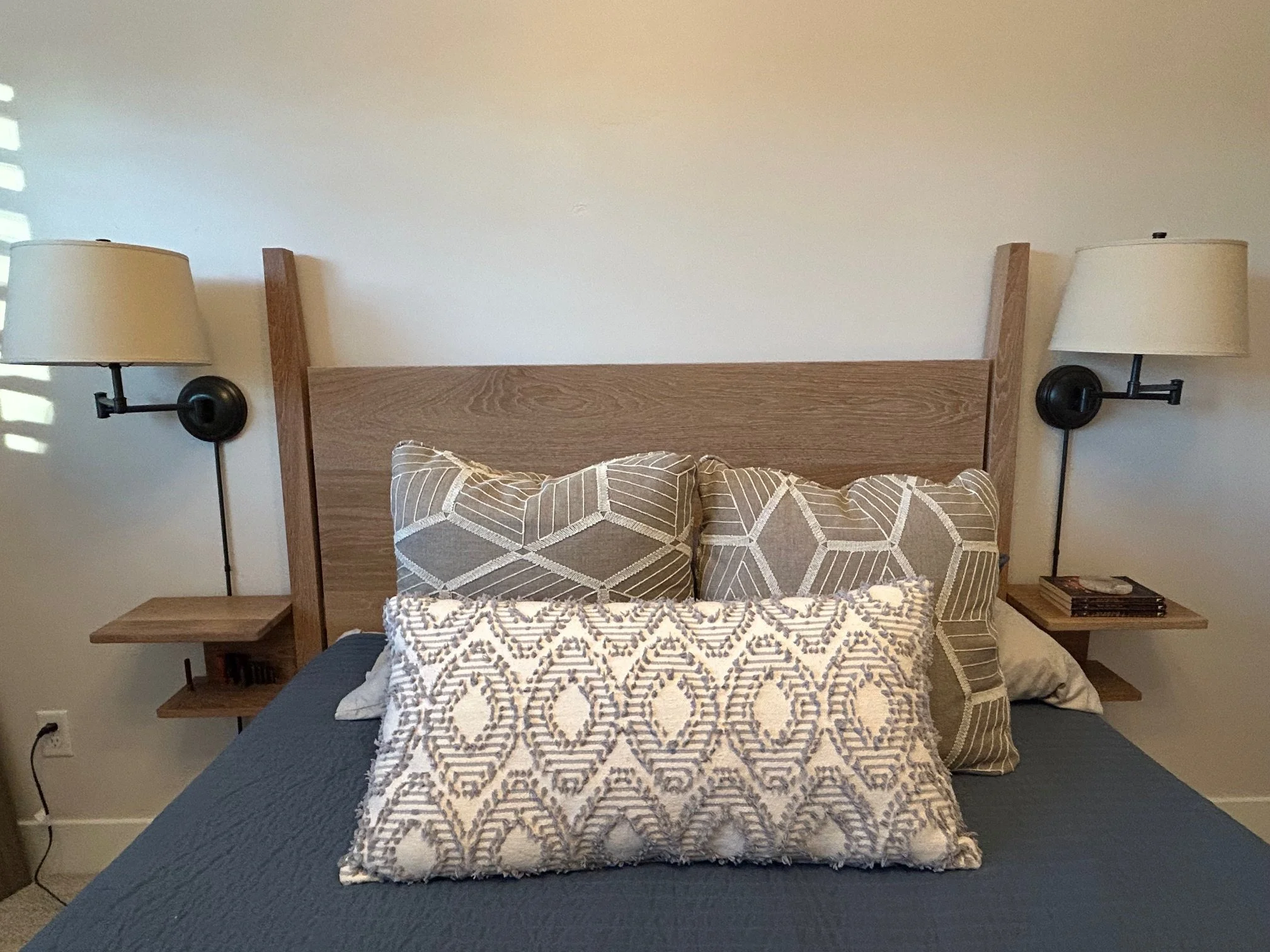 A bedroom with a wooden bed frame, two pillows with patterned covers, two black wall-mounted lamps with white shades, and small wooden nightstands on each side of the bed.